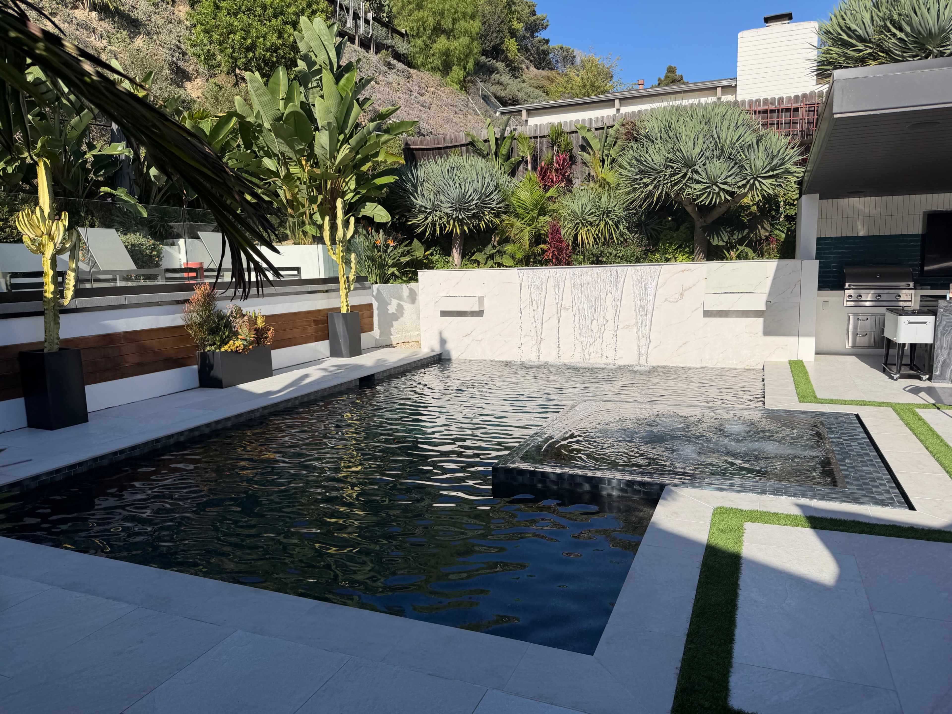 The image shows a modern backyard with a swimming pool, a water feature, tropical plants, and patio furniture under a clear blue sky.