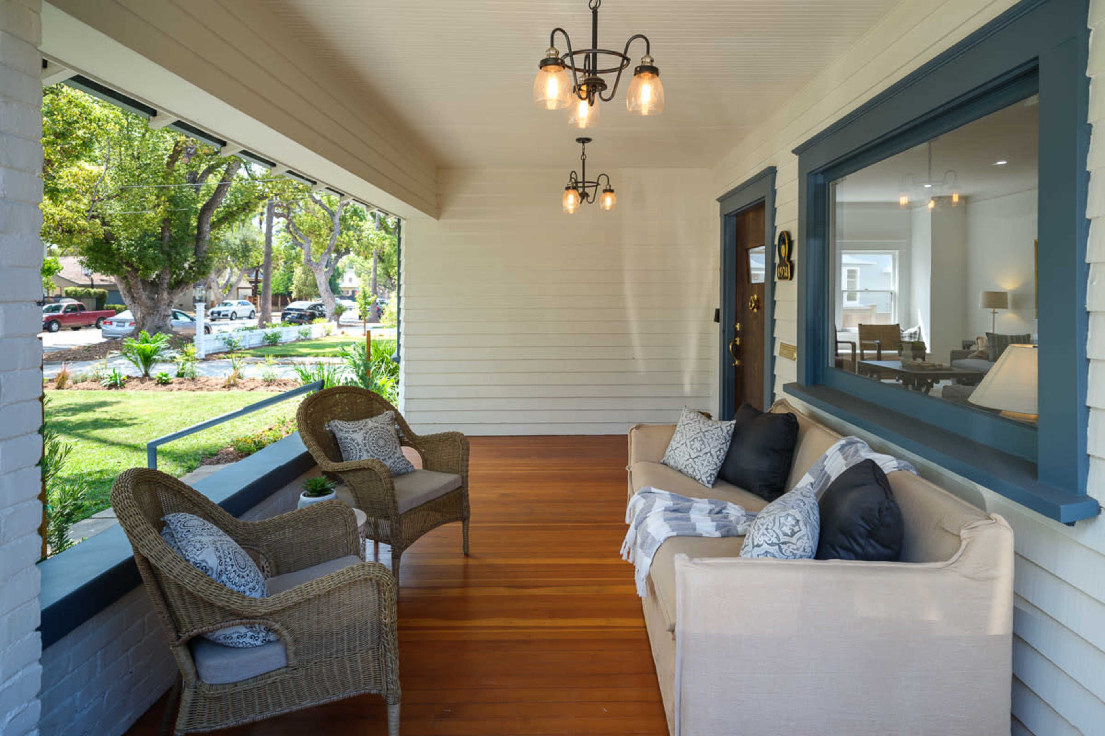 The image shows a porch featuring two wicker chairs and a sofa adorned with decorative pillows, all set on polished wooden flooring under hanging lights.
