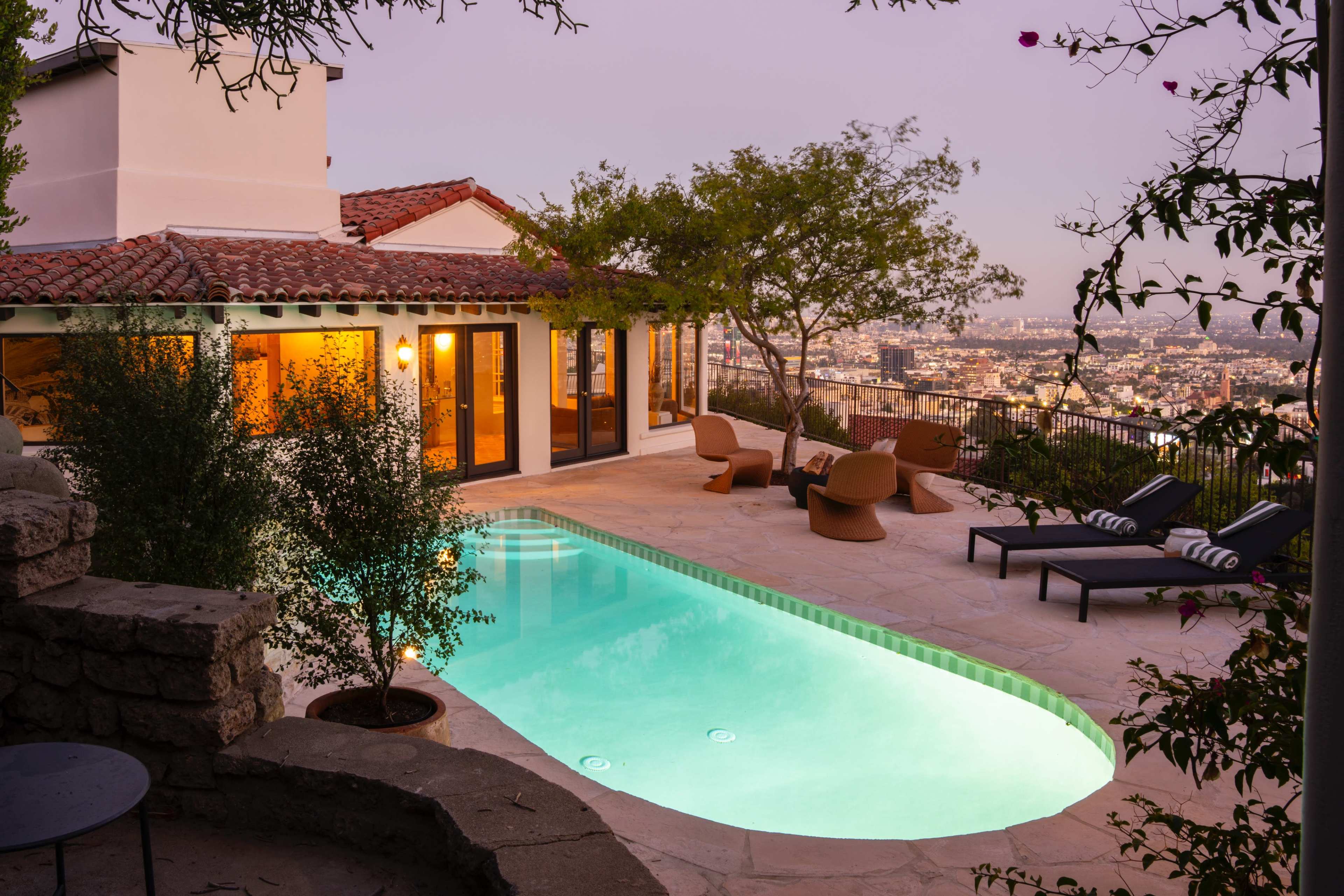The image shows a clear swimming pool surrounded by a stone patio and lounge chairs, with a house nearby and a city skyline visible in the distance at twilight.