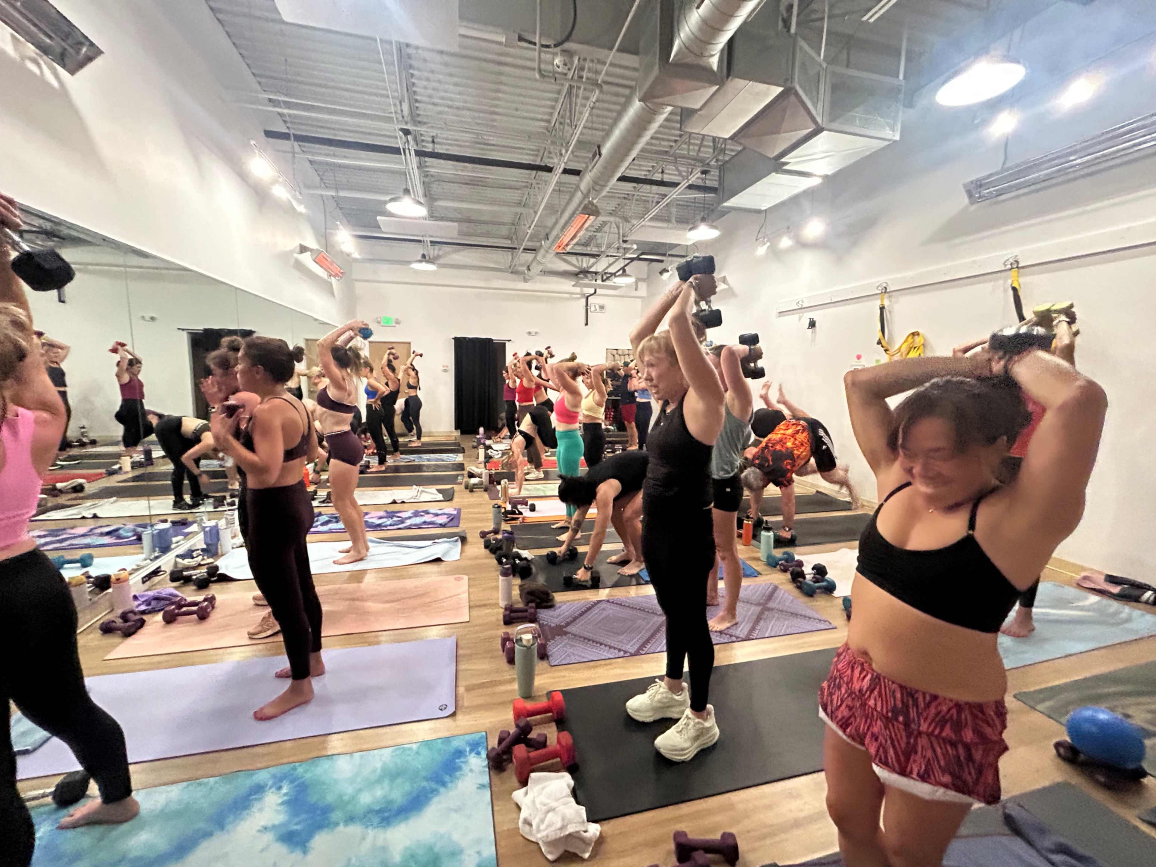 A large group of people is participating in a workout class, lifting weights and exercising on mats in a bright, spacious studio.
