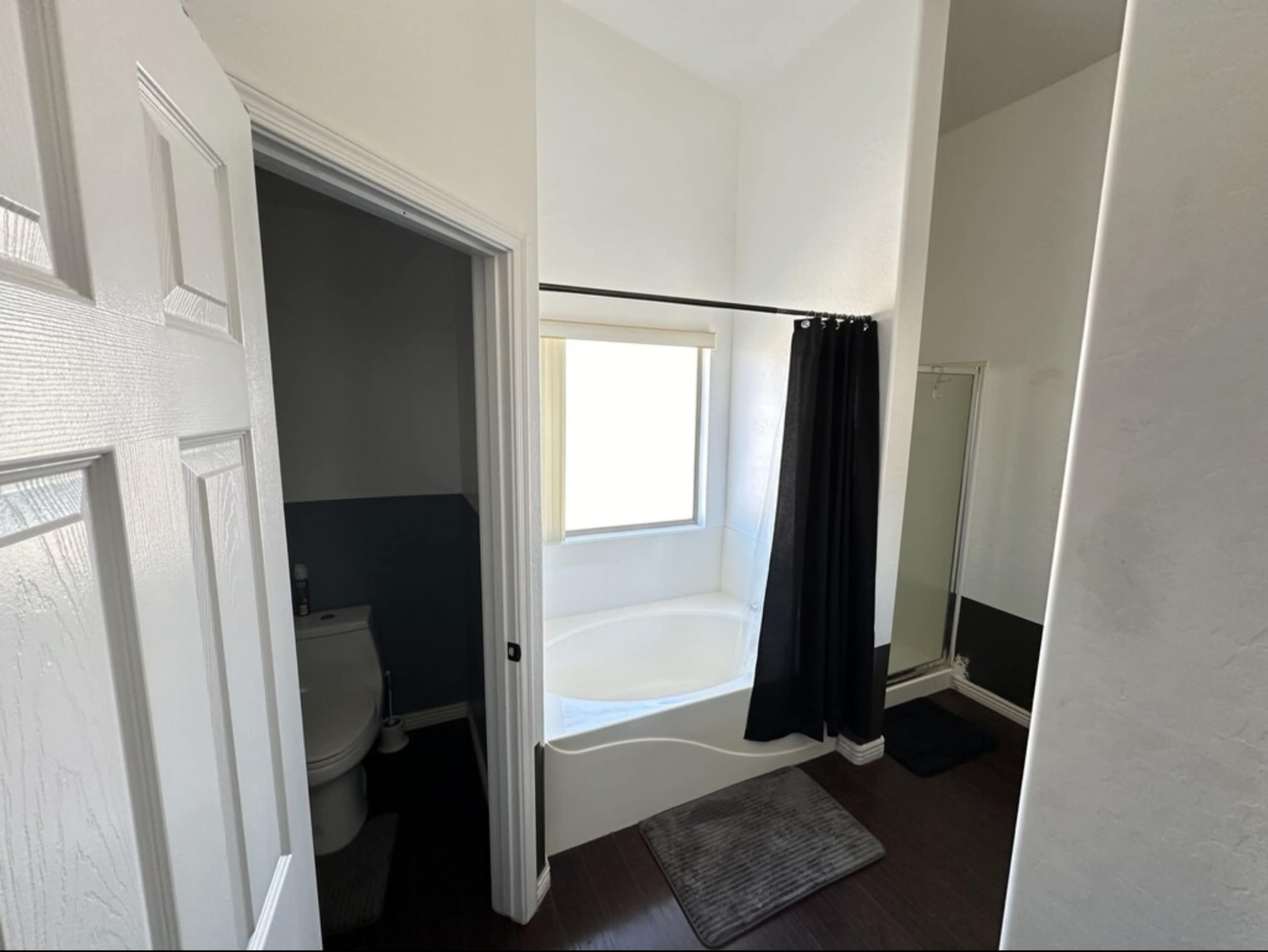 The image shows a bathroom featuring a corner bathtub, a shower area, a toilet, and a window with natural light.