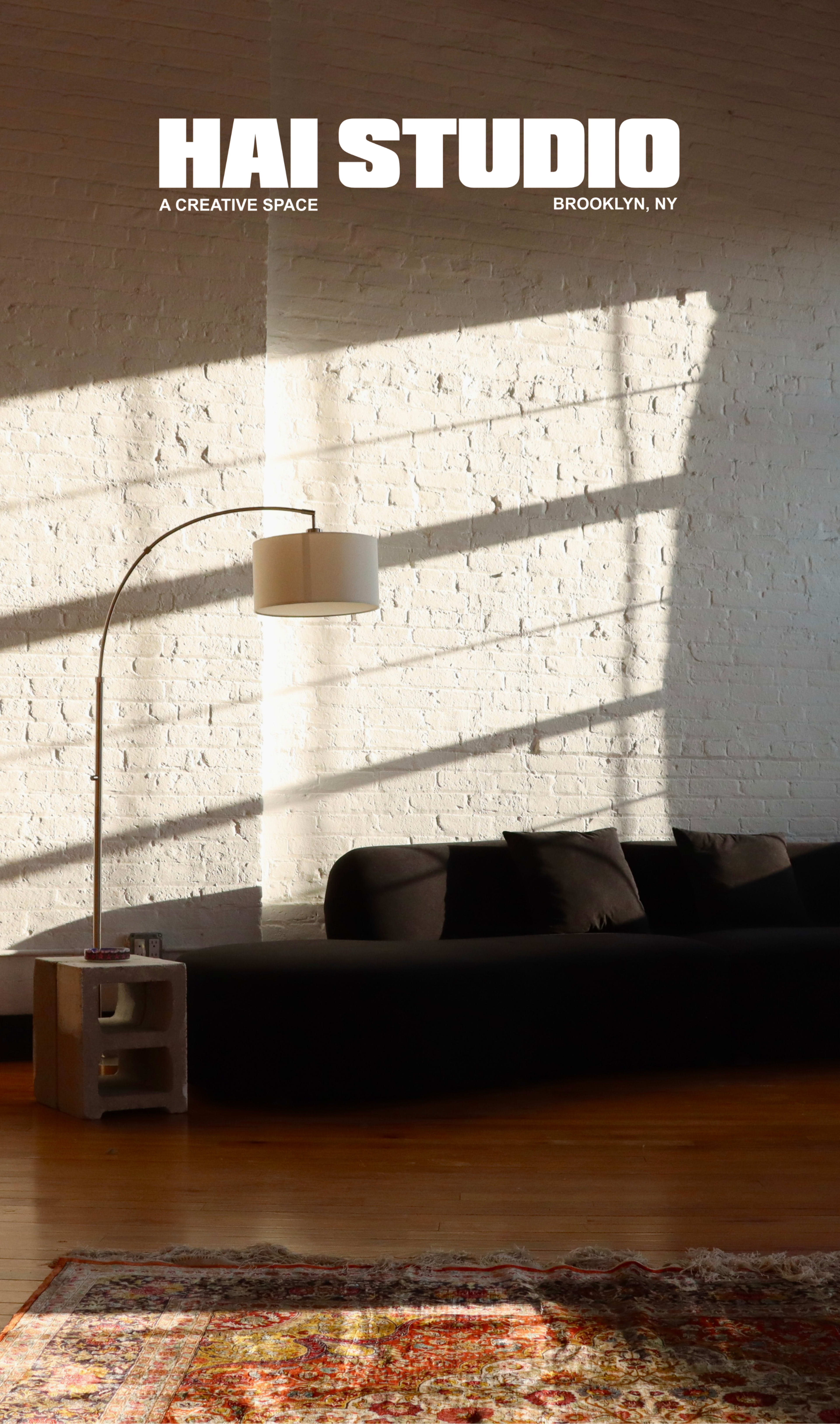 The image depicts a minimalistic studio space with a black sofa, a floor lamp, and sunlight casting shadows on a white brick wall.