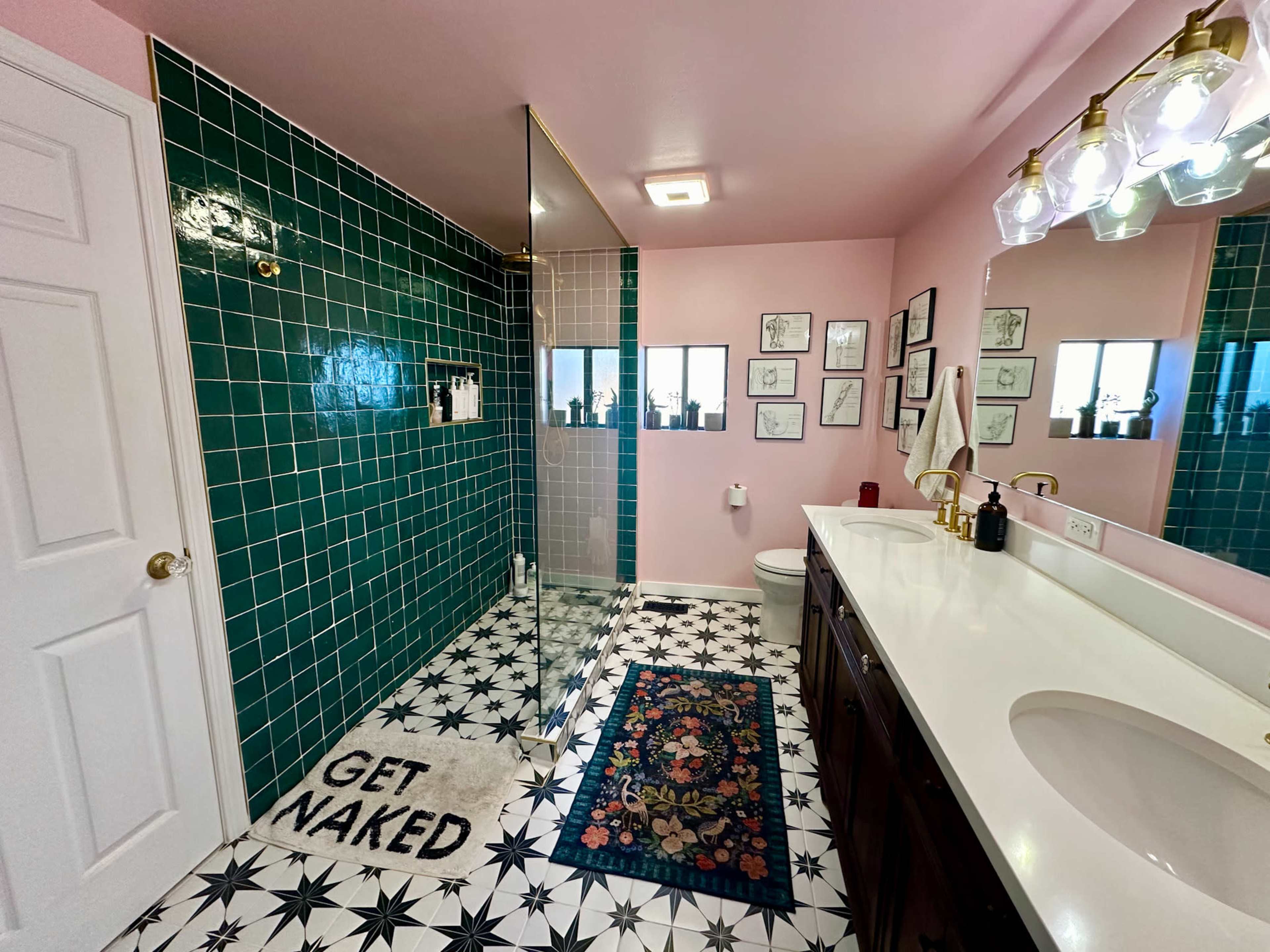 The bathroom features green tiled walls, a glass shower, a white countertop with dark wood cabinets, and a patterned floor with a decorative rug.
