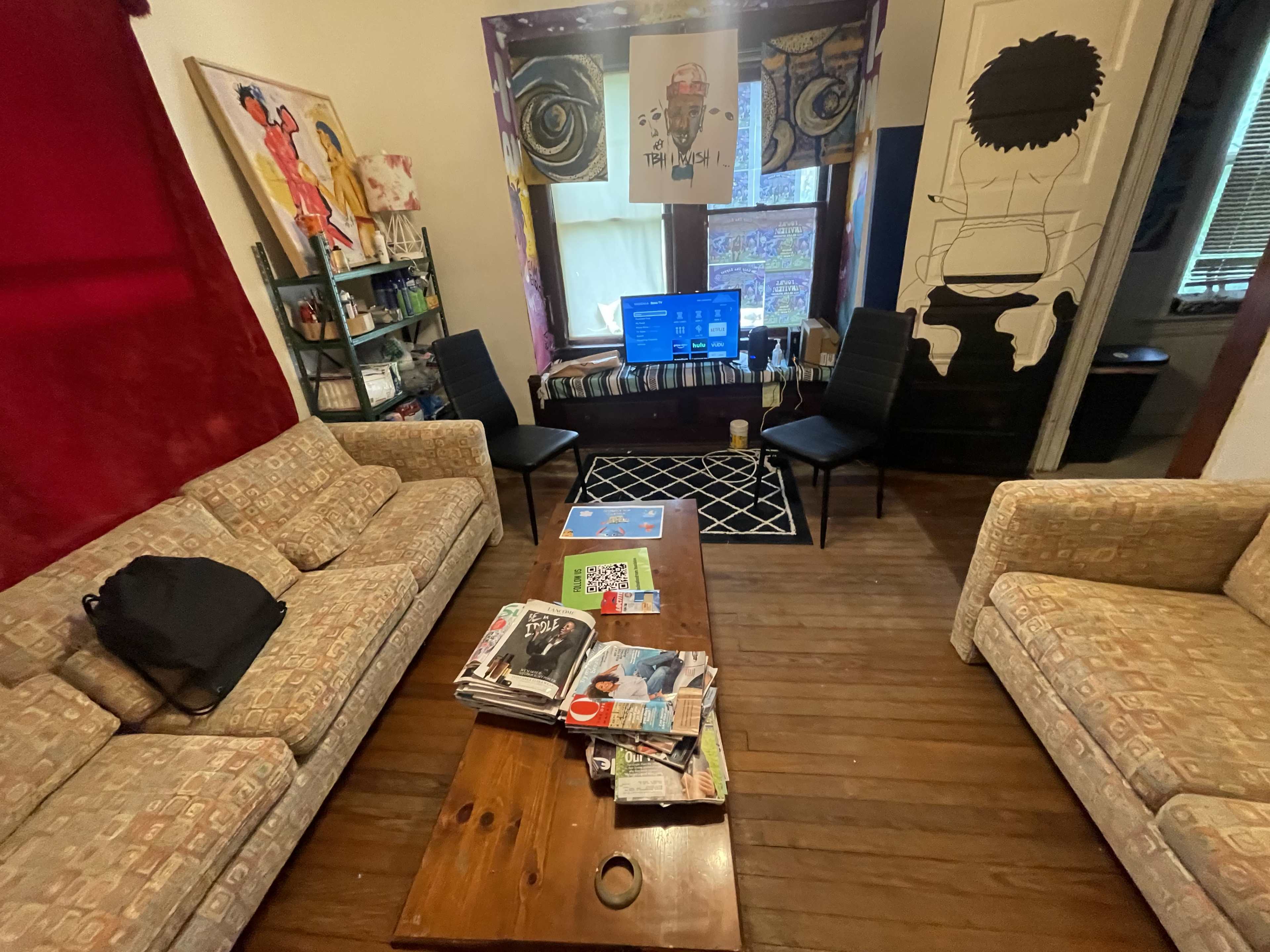 The image shows a living room with two patterned sofas facing a wooden coffee table, a television on a stand, and a mural on the wall.