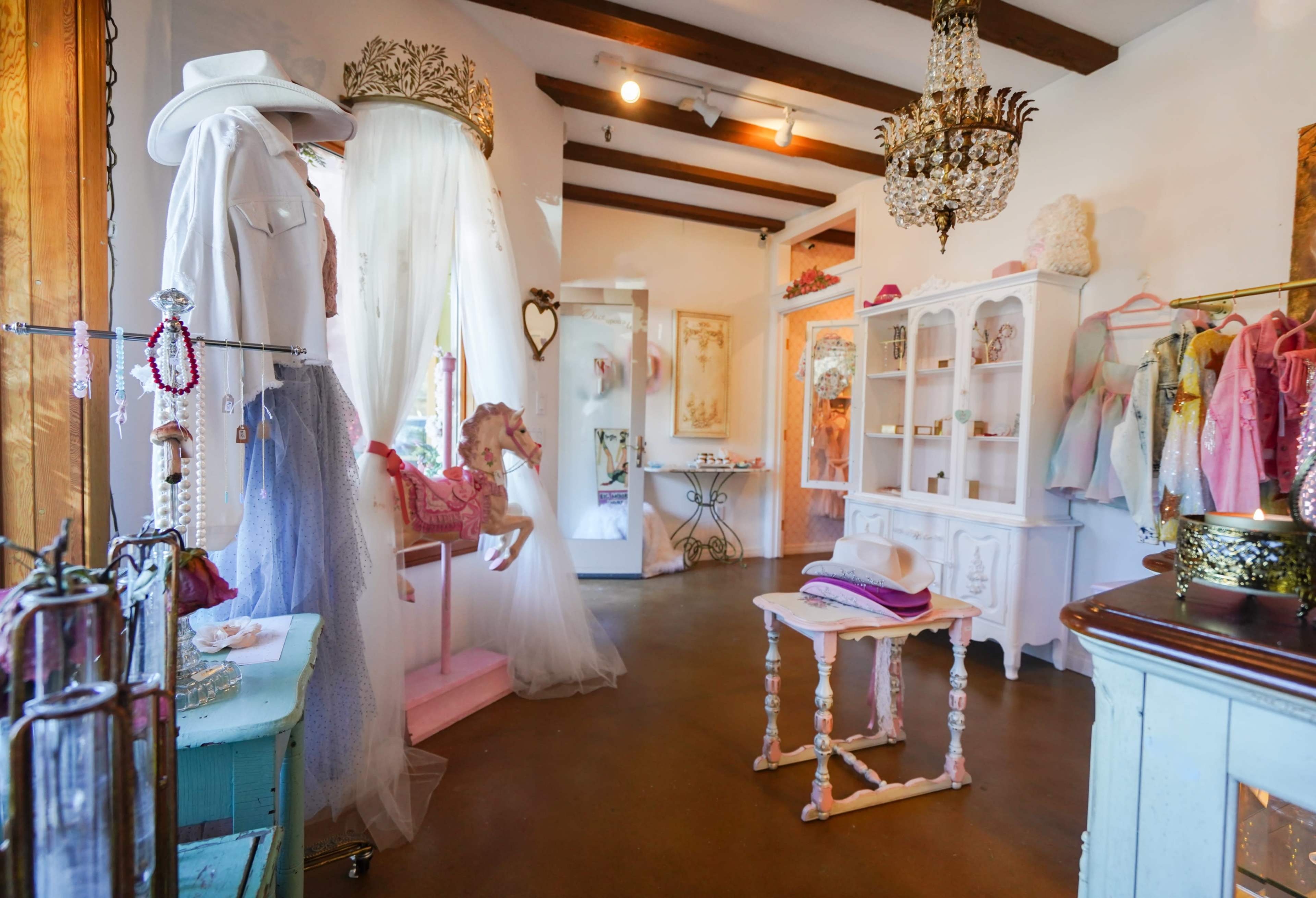 A boutique interior featuring a mannequin in white attire, a pink horse display, a glass cabinet, and vintage decor, with a chandelier overhead.
