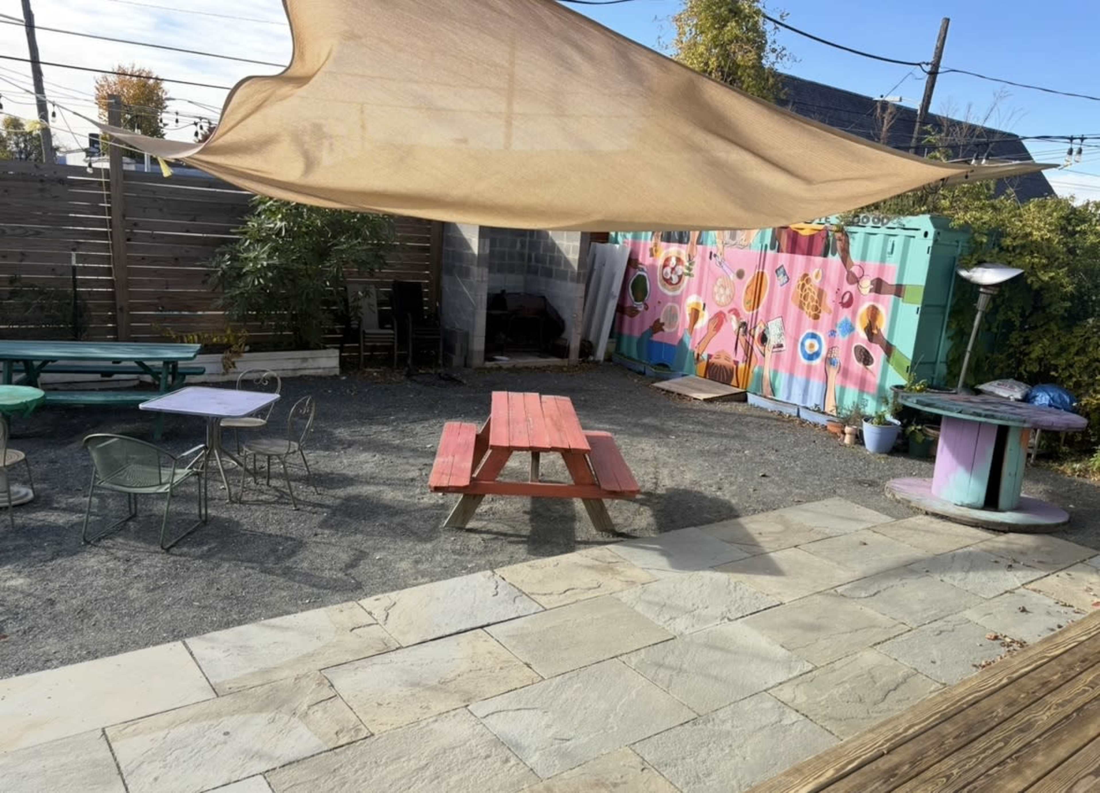 The image shows an outdoor seating area with various tables and chairs, a picnic table, and a colorful wall mural, all covered partially by a large shade sail.