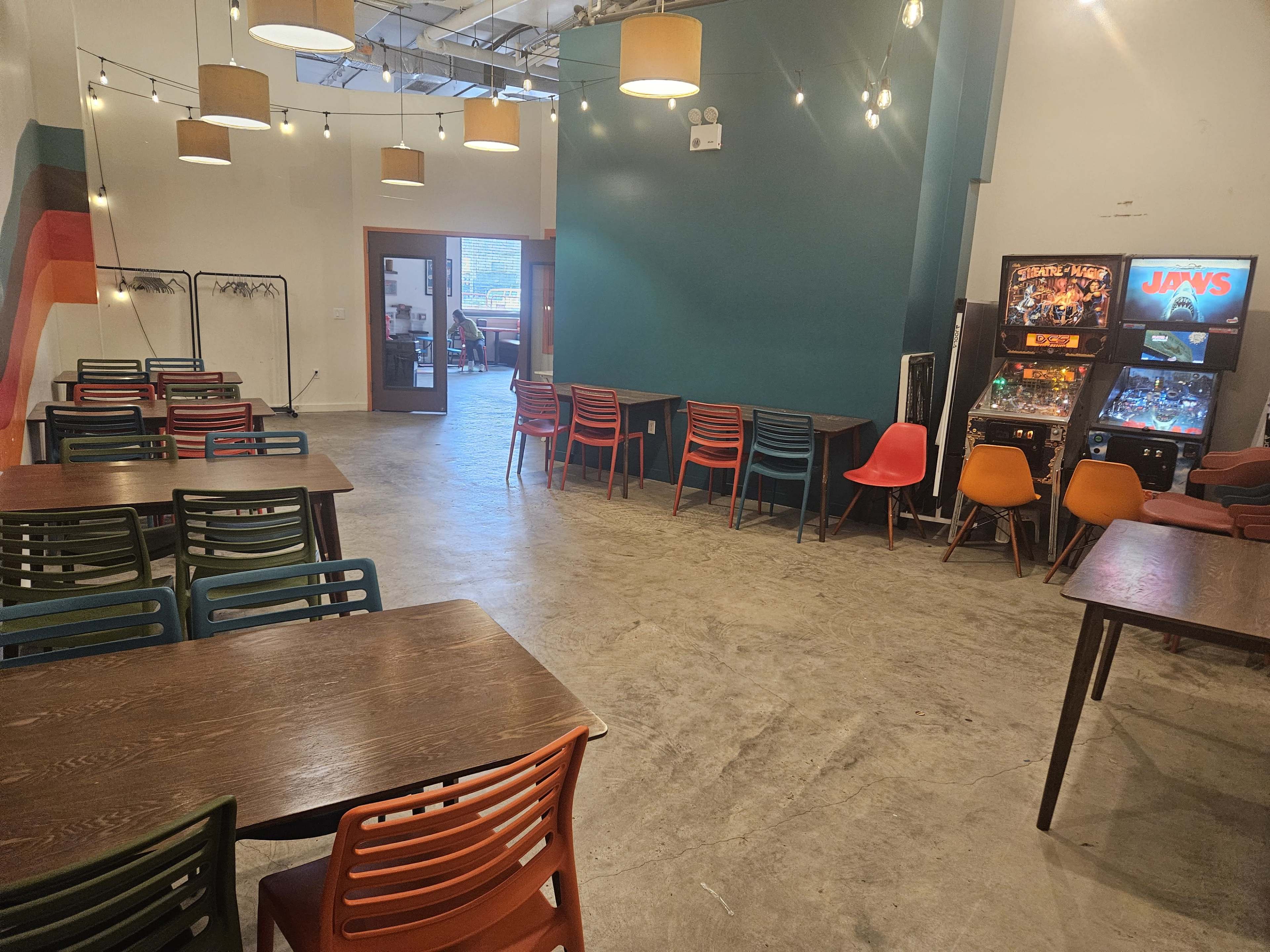 The image shows a spacious indoor area with colorful chairs and tables arranged for seating, featuring a pinball machine and a utility rack in the background.