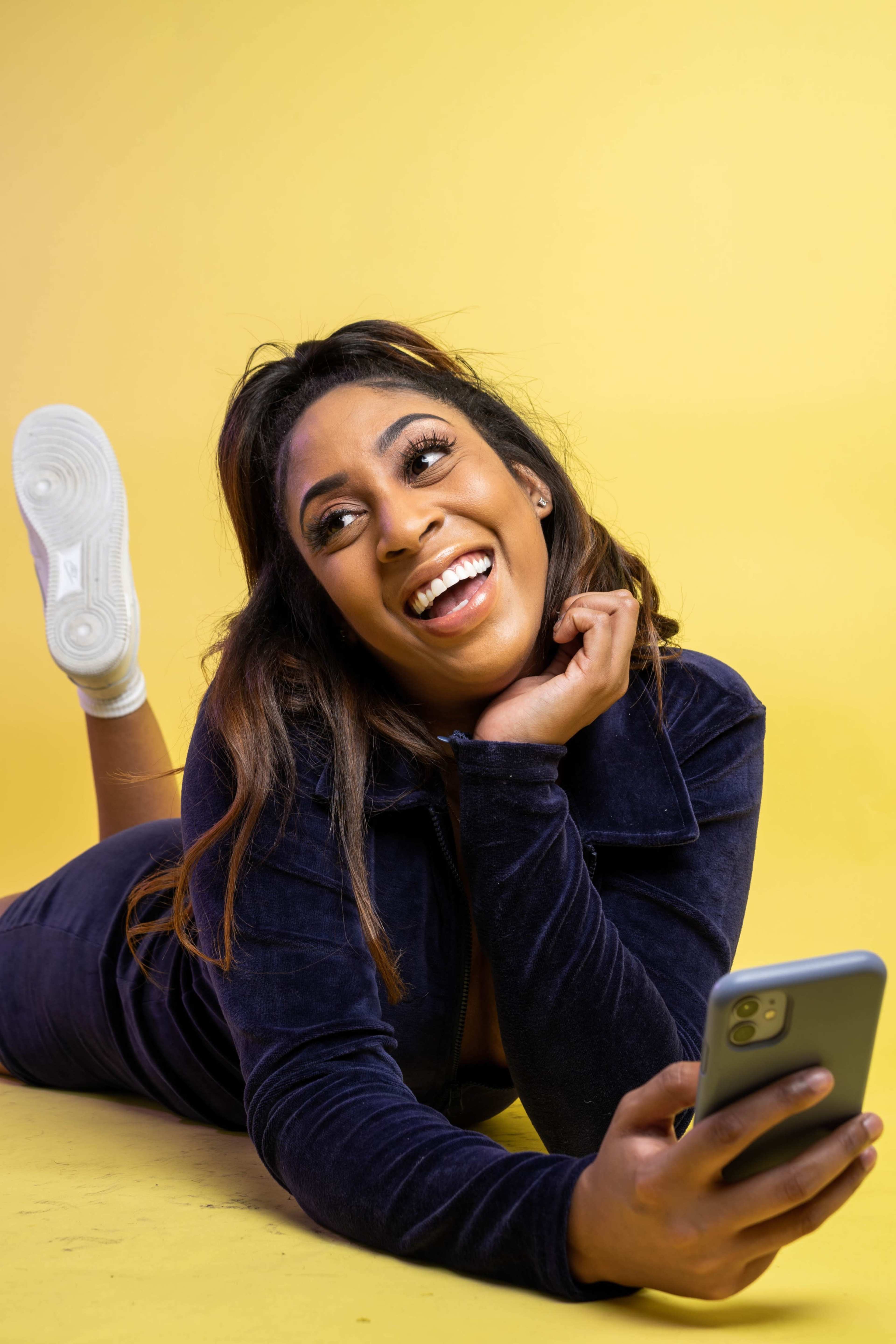 A woman lies on her stomach on a yellow background, smiling while holding a smartphone.