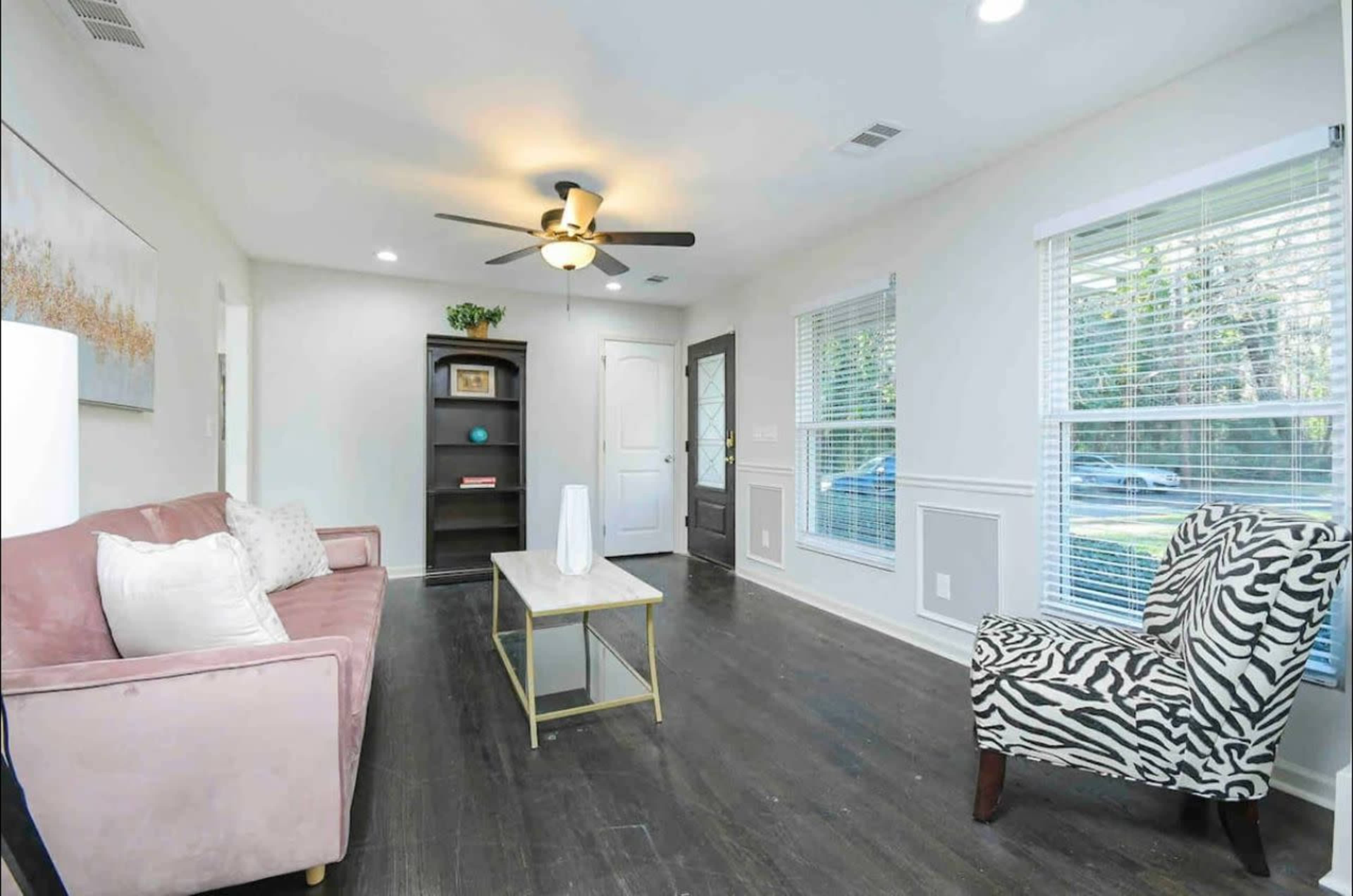 A living room features a pink sofa, a zebra print chair, a coffee table, and a bookcase against a wall with large windows letting in natural light.