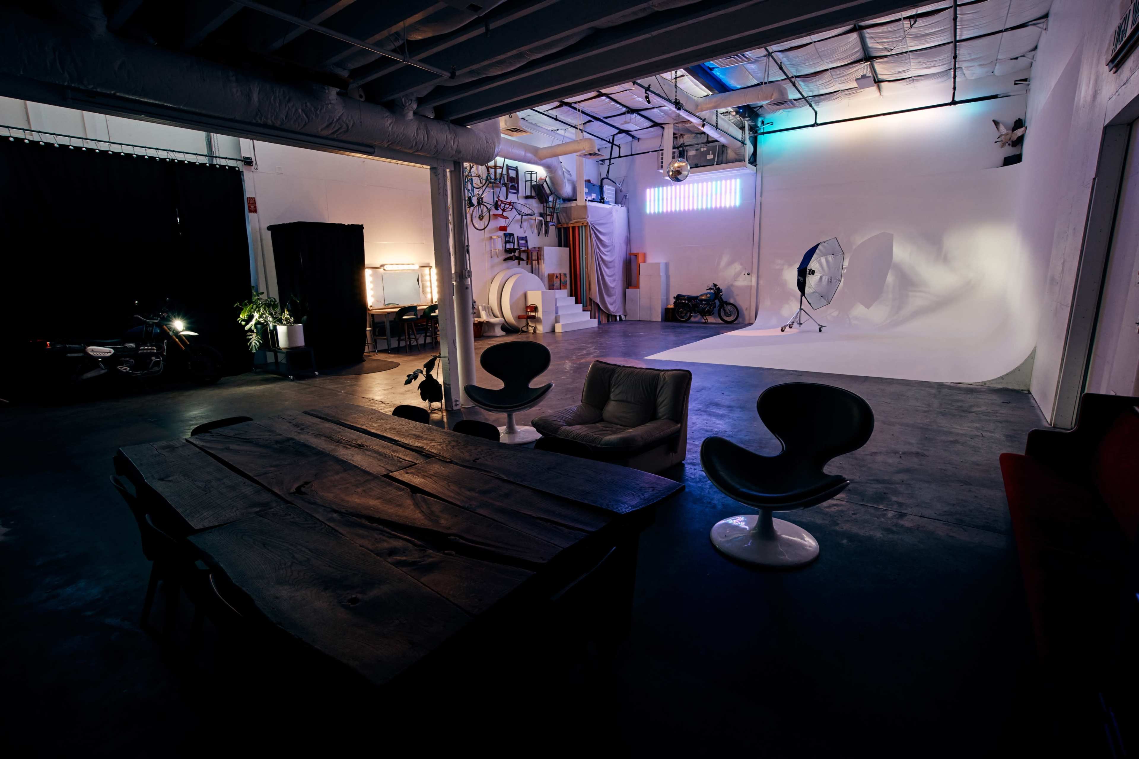 An industrial-style studio space featuring a large wooden table, several chairs, and a photography backdrop lit with soft lighting.