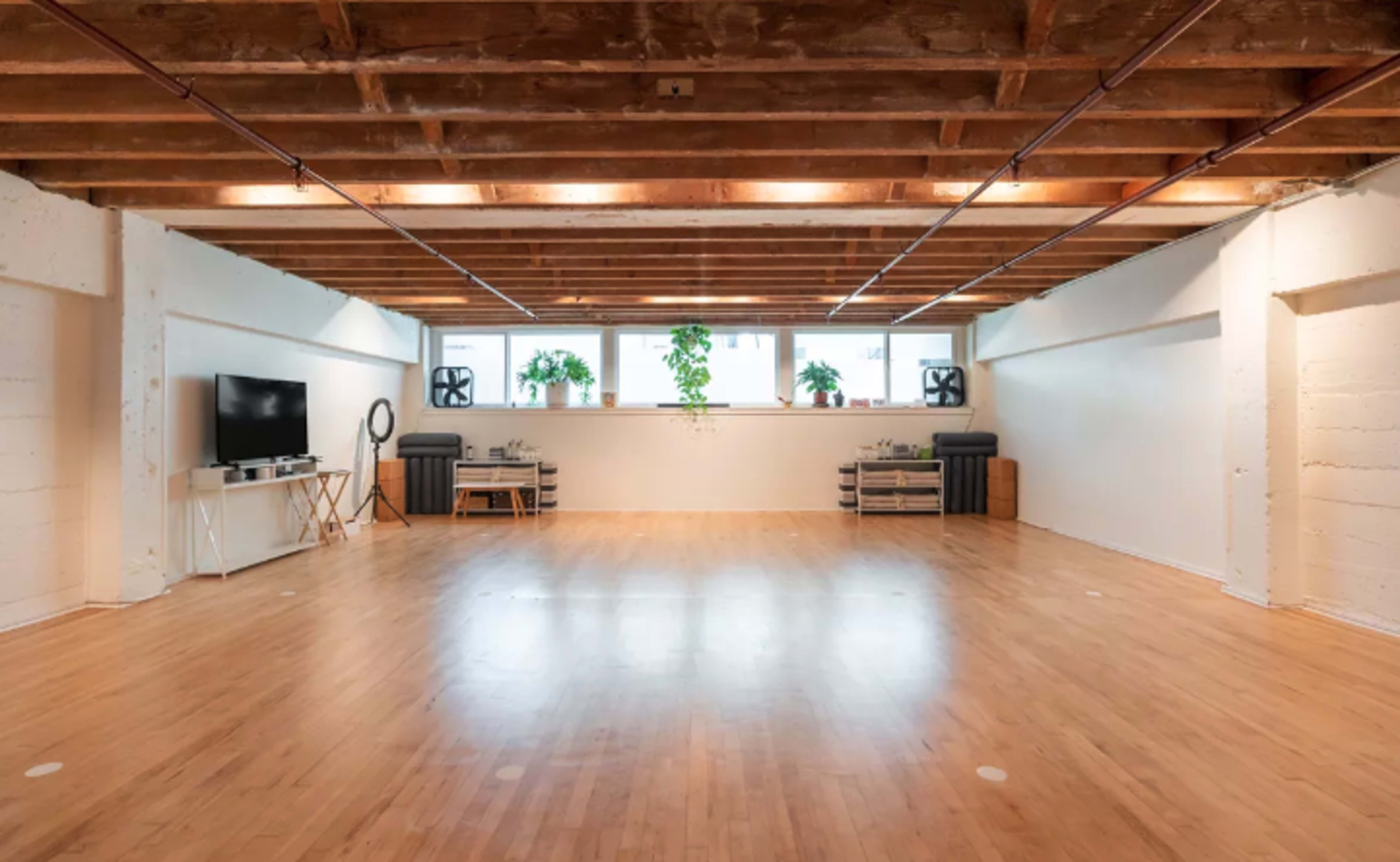 A spacious, empty room with hardwood flooring, exposed wooden beams, large windows, and potted plants.