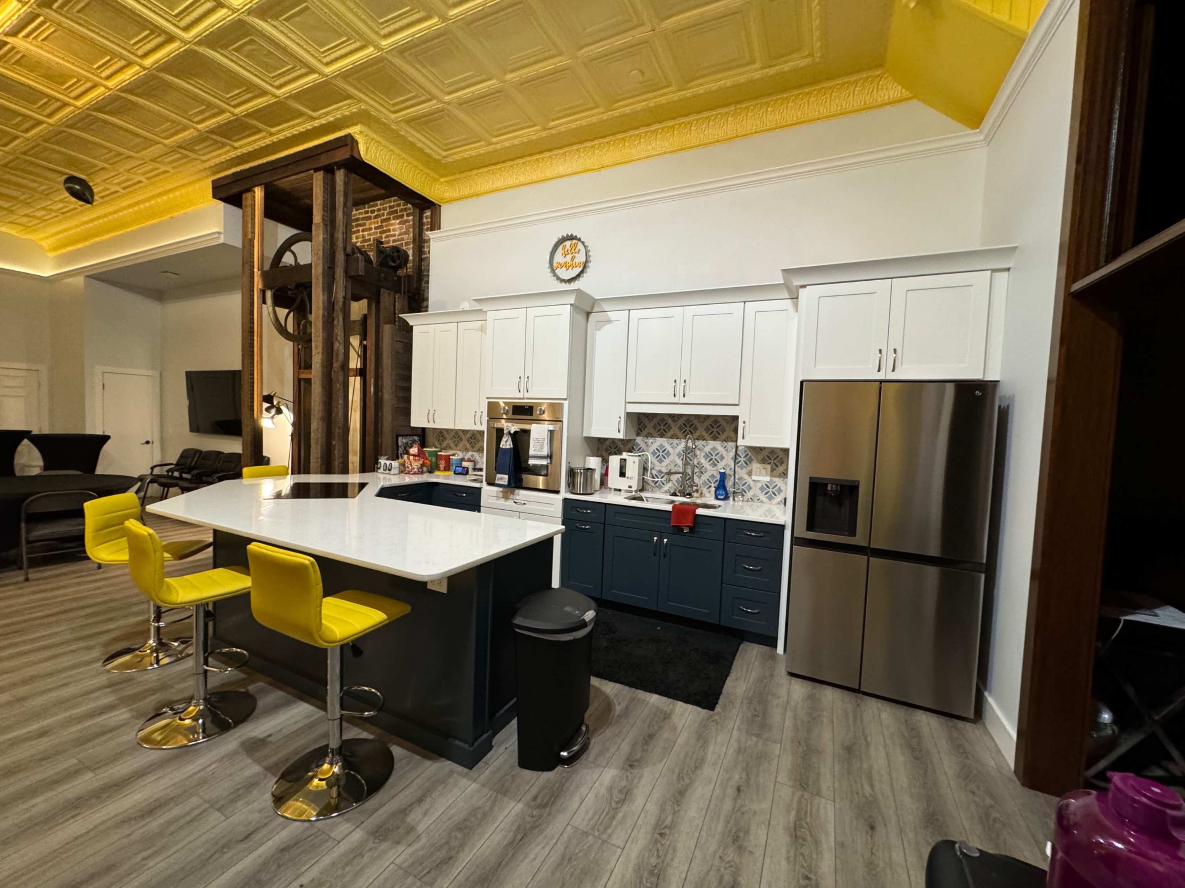 The image shows a modern kitchen with white cabinets, a gray island topped with a white countertop, yellow bar stools, and stainless steel appliances.