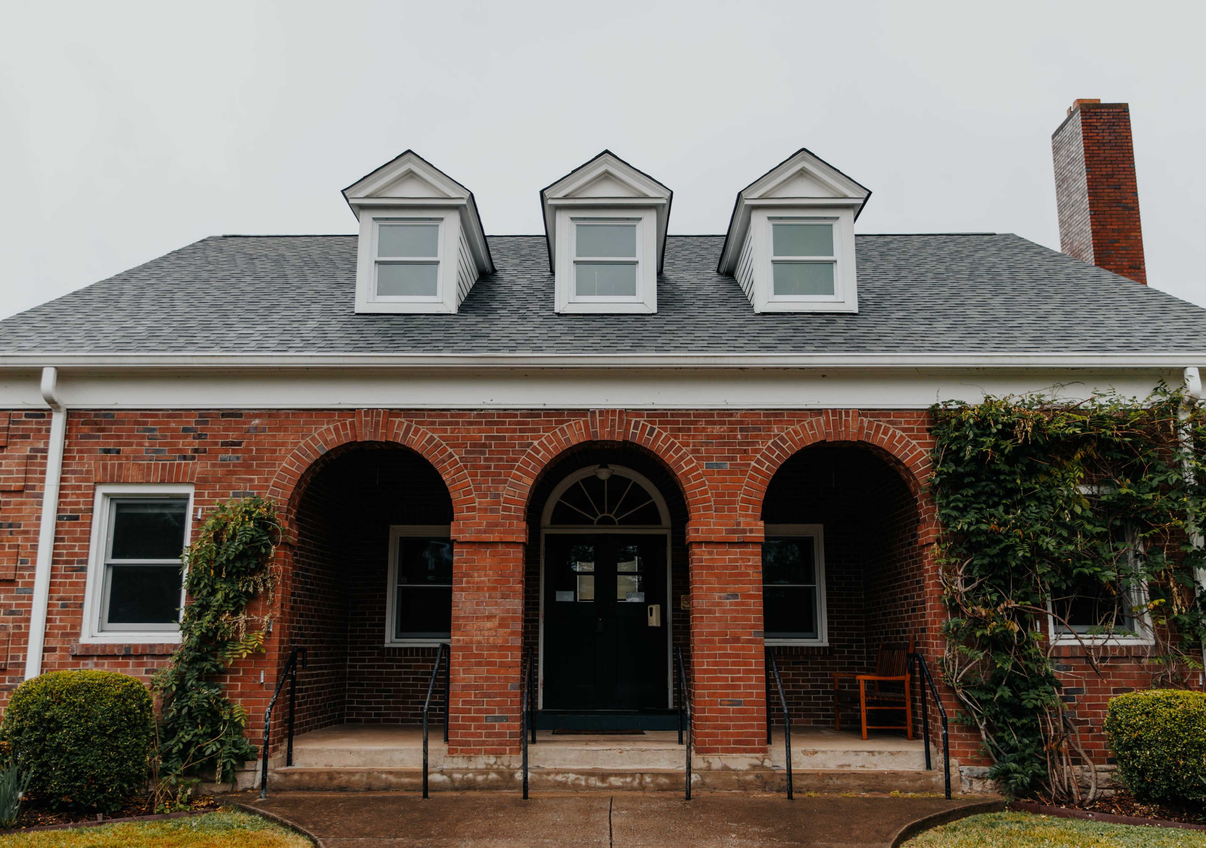 The image shows a brick building with three dormer windows and arched entrances, flanked by small hedges on either side.