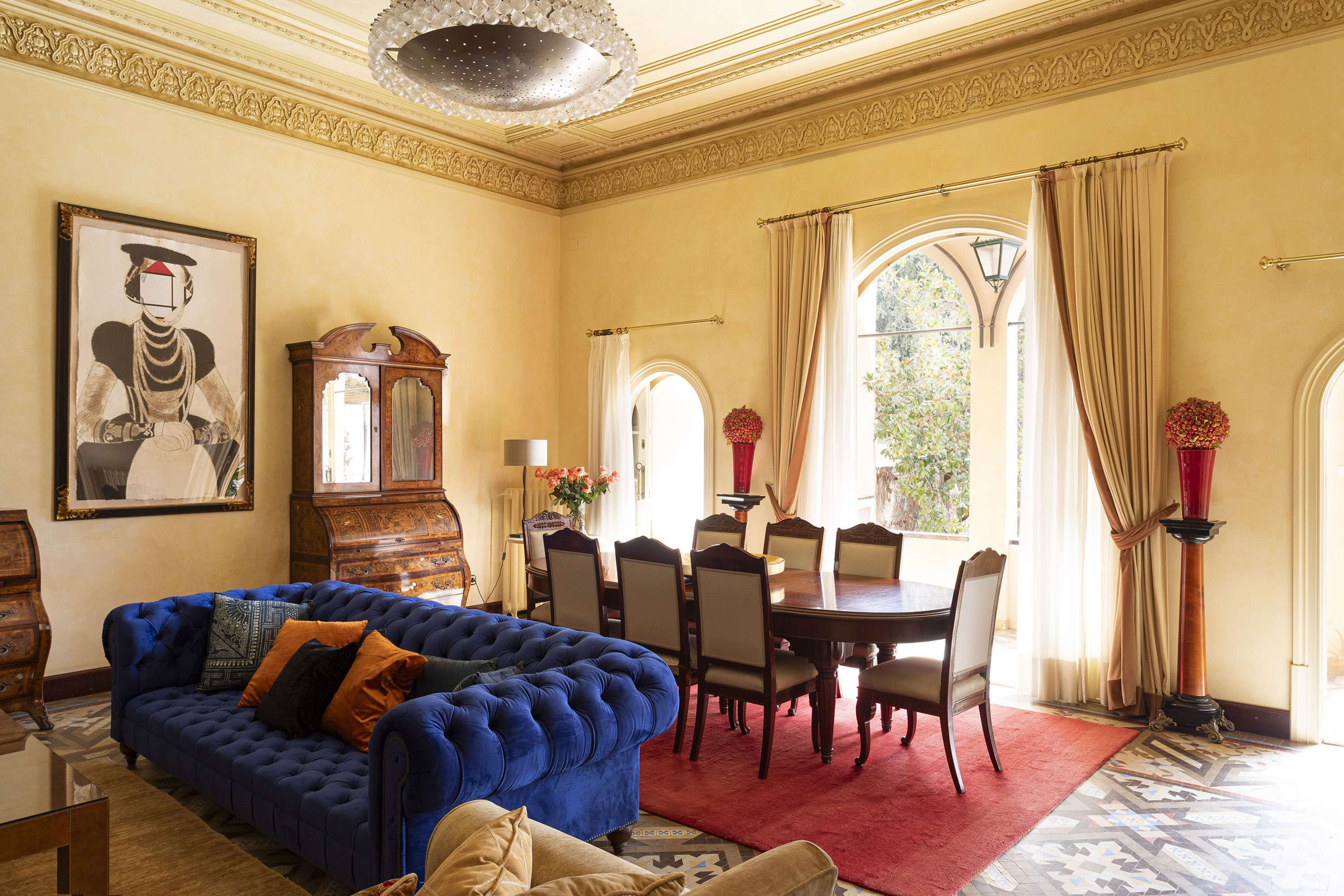 A spacious room with a blue velvet sofa, a wooden dining table surrounded by chairs, and large windows framed by drapes.