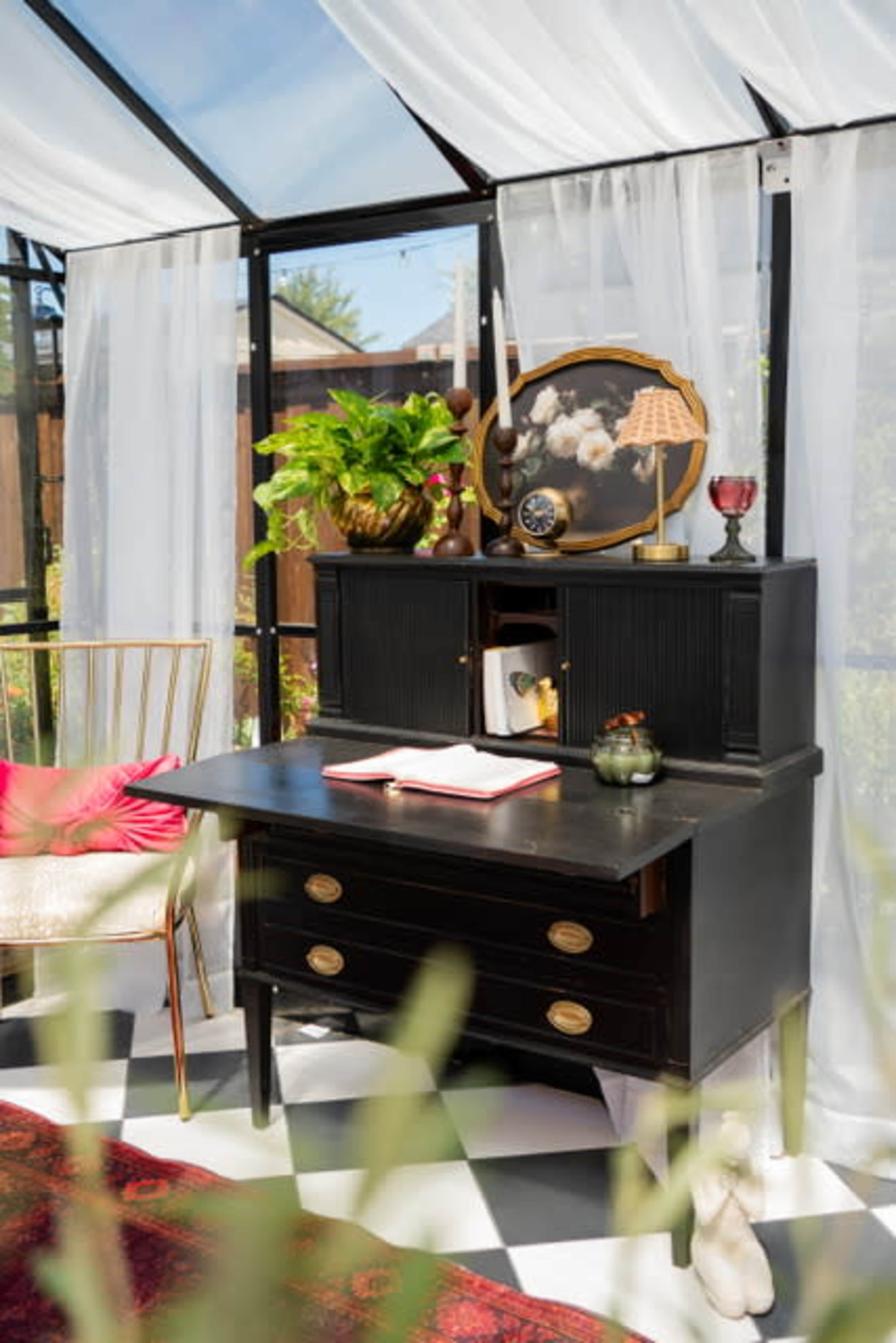 A black vintage desk with a hutch is positioned in a sunlit glass structure, adorned with decorative items and surrounded by greenery.