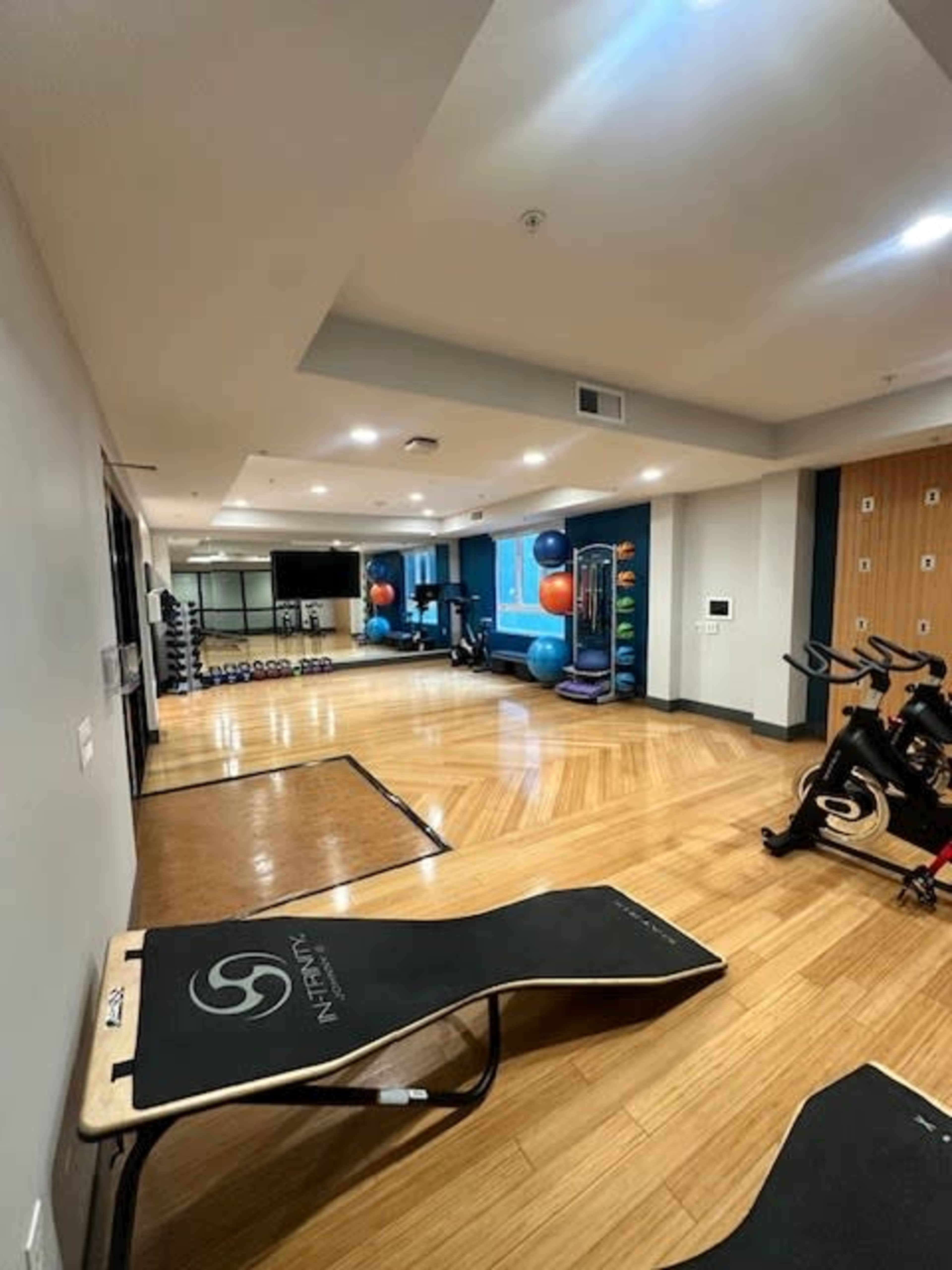 The image shows a fitness area with exercise equipment, including weights, a stationary bike, and yoga balls, arranged in a spacious room with wooden flooring.