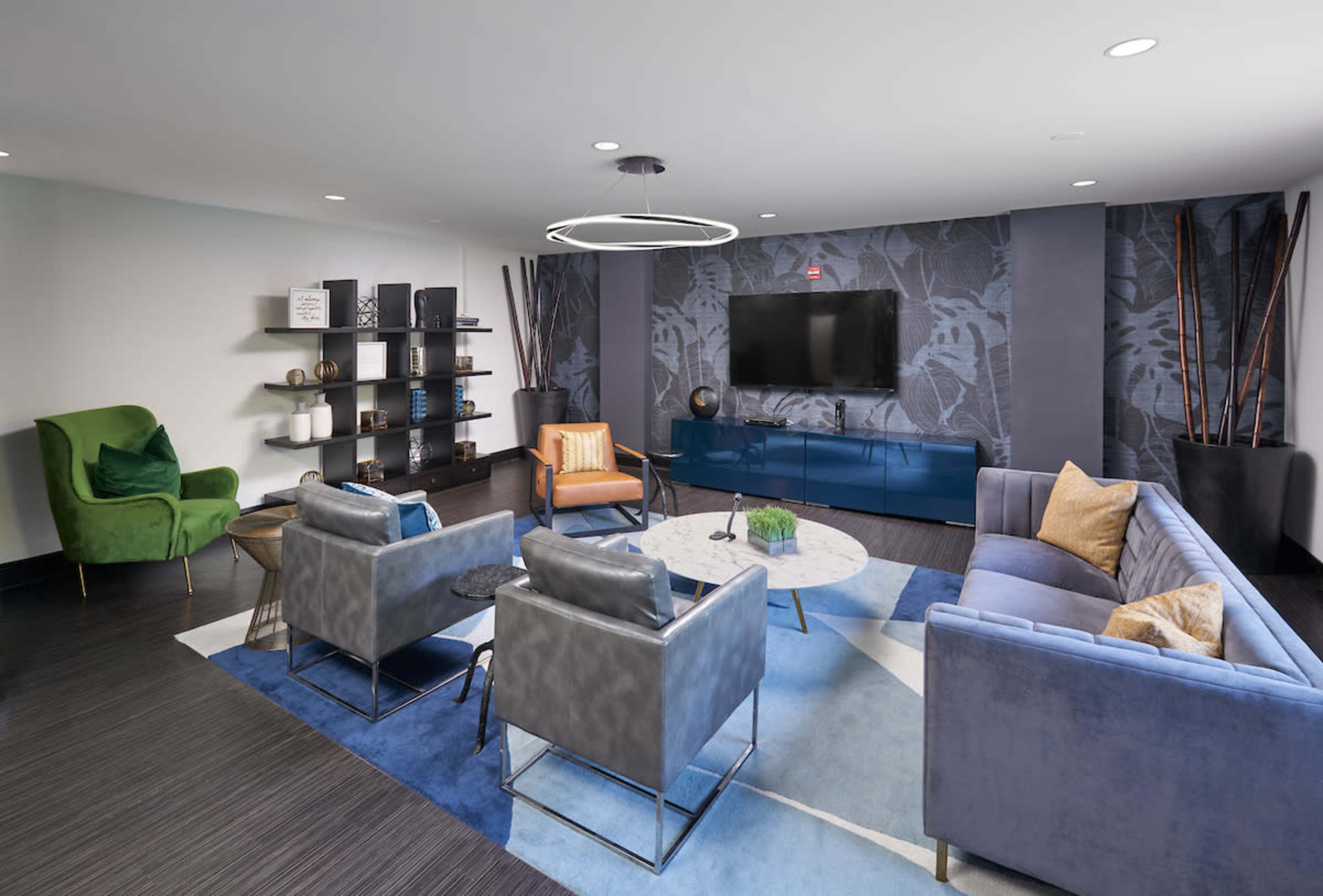 A modern living room with a blue and gray color scheme, featuring a television mounted on the wall, a seating area with various couches and chairs, and a bookshelf displaying decorative items.