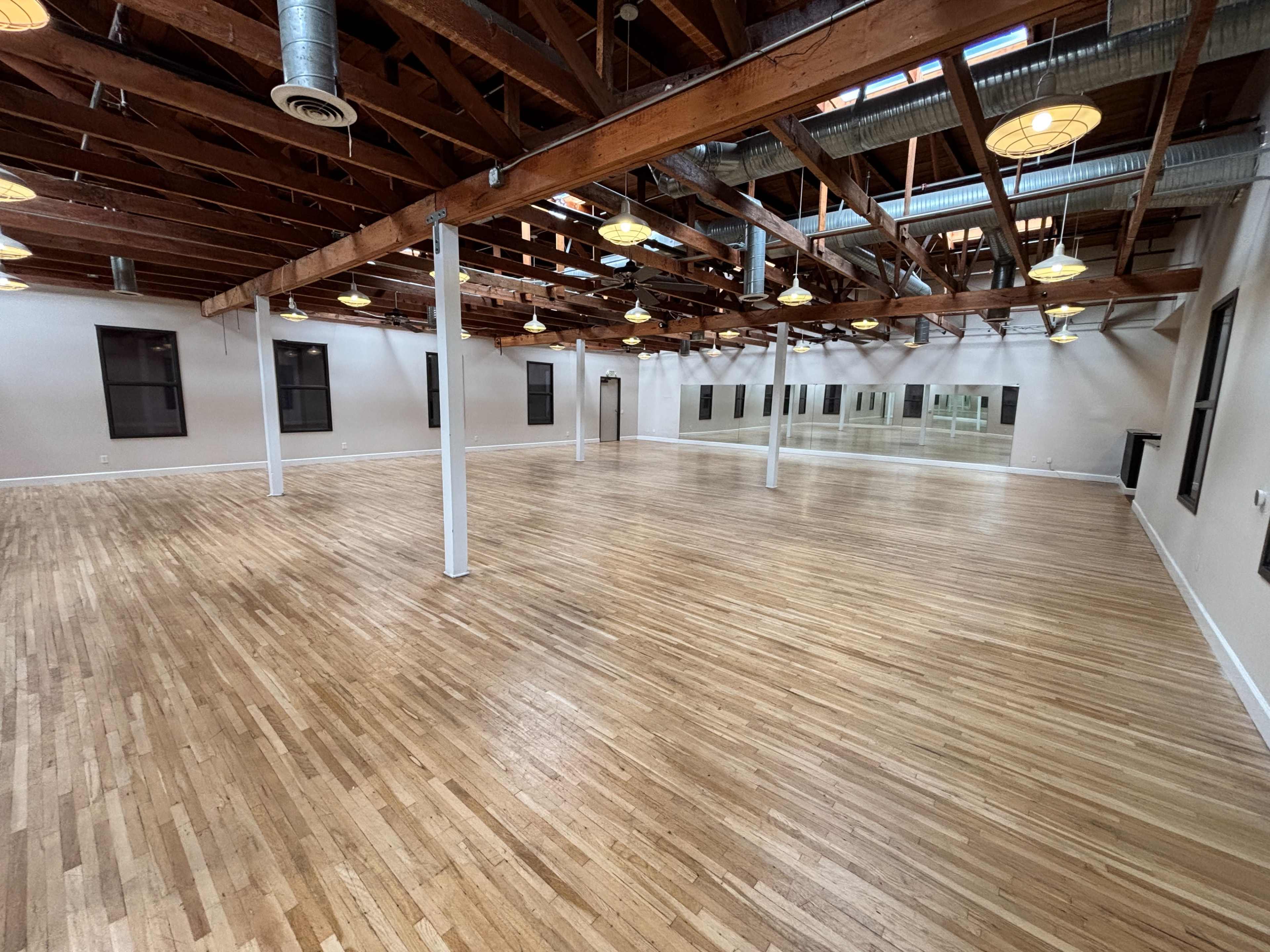 The image shows an empty room with polished wooden floors, exposed wooden beams, and large windows, creating a spacious, well-lit environment.