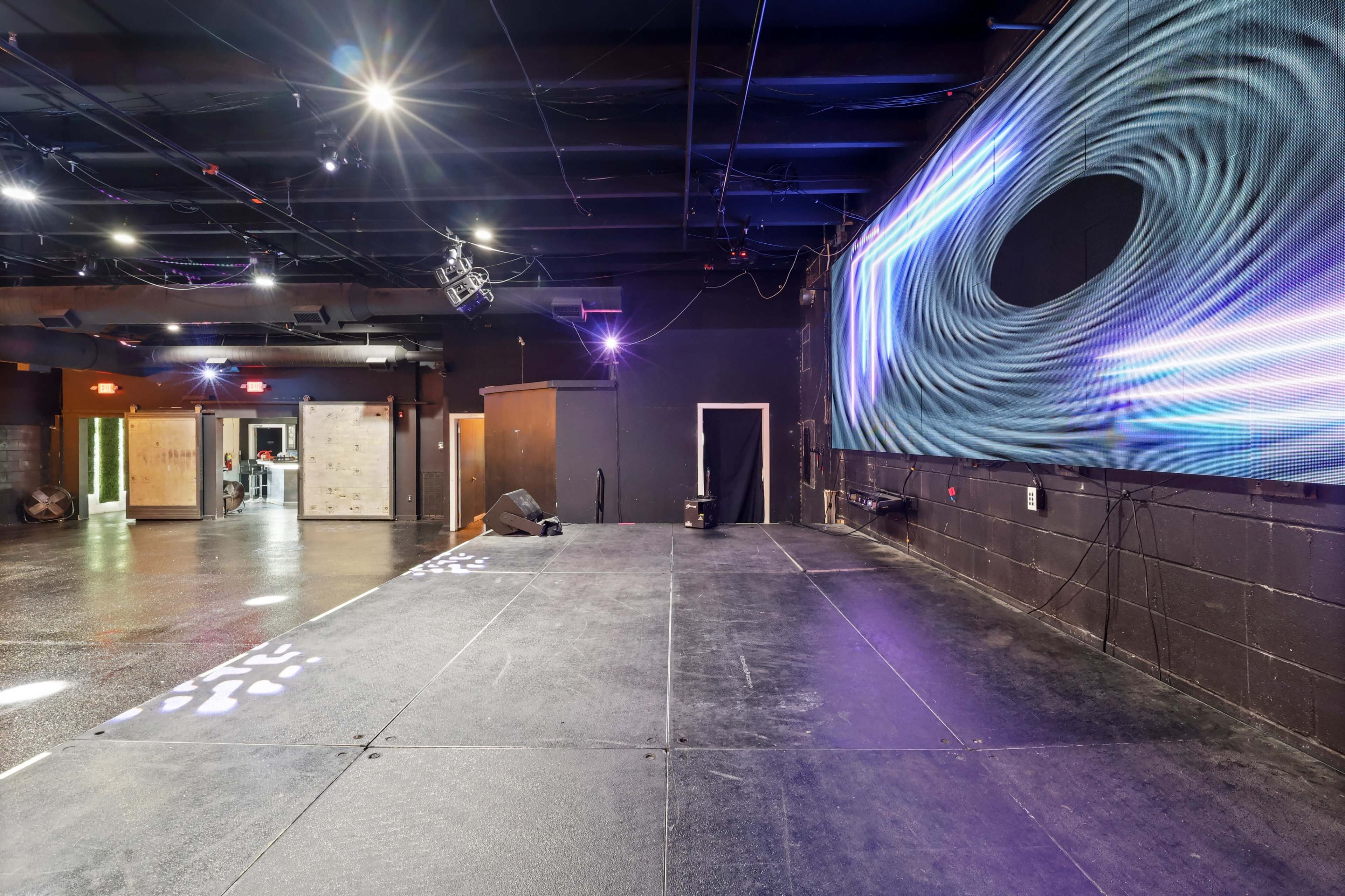 The image shows a spacious performance stage area with a large, illuminated screen on one wall and various entranceways visible in the background.
