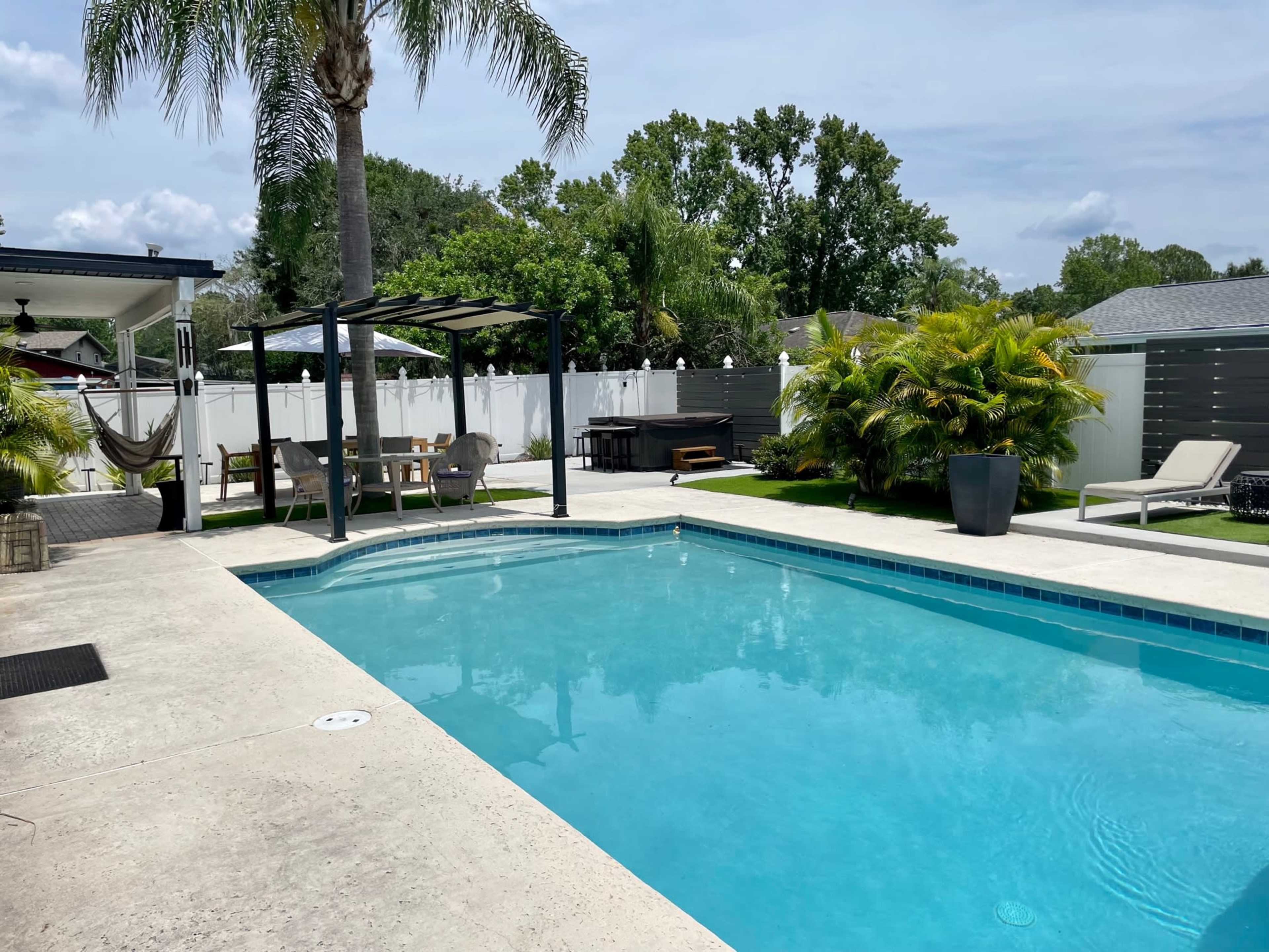 A clear blue swimming pool surrounded by a concrete patio, a palm tree, outdoor furniture, and a lush garden area.