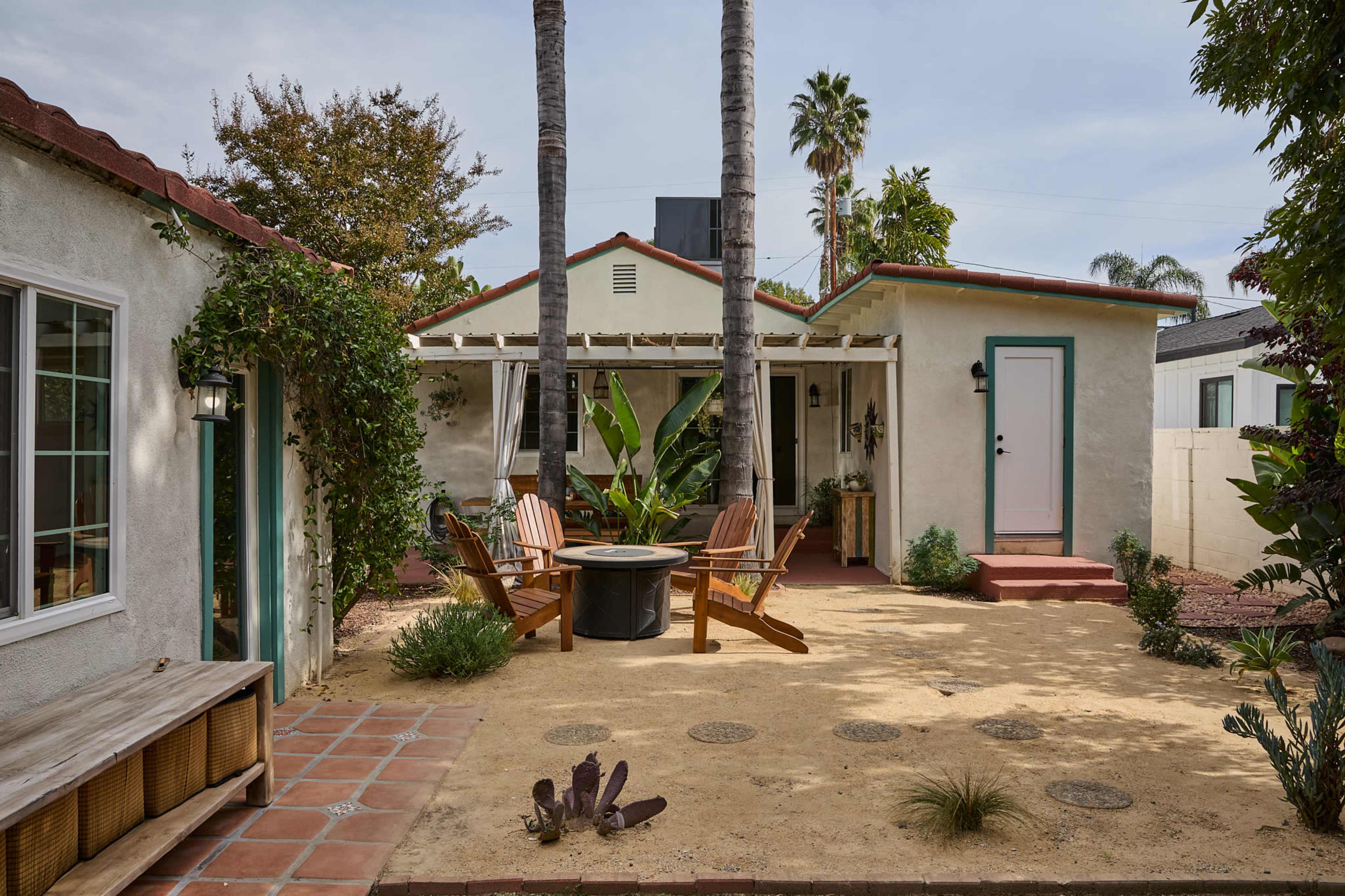 The image shows a small courtyard with wooden chairs arranged around a fire pit, framed by palm trees and surrounded by greenery.