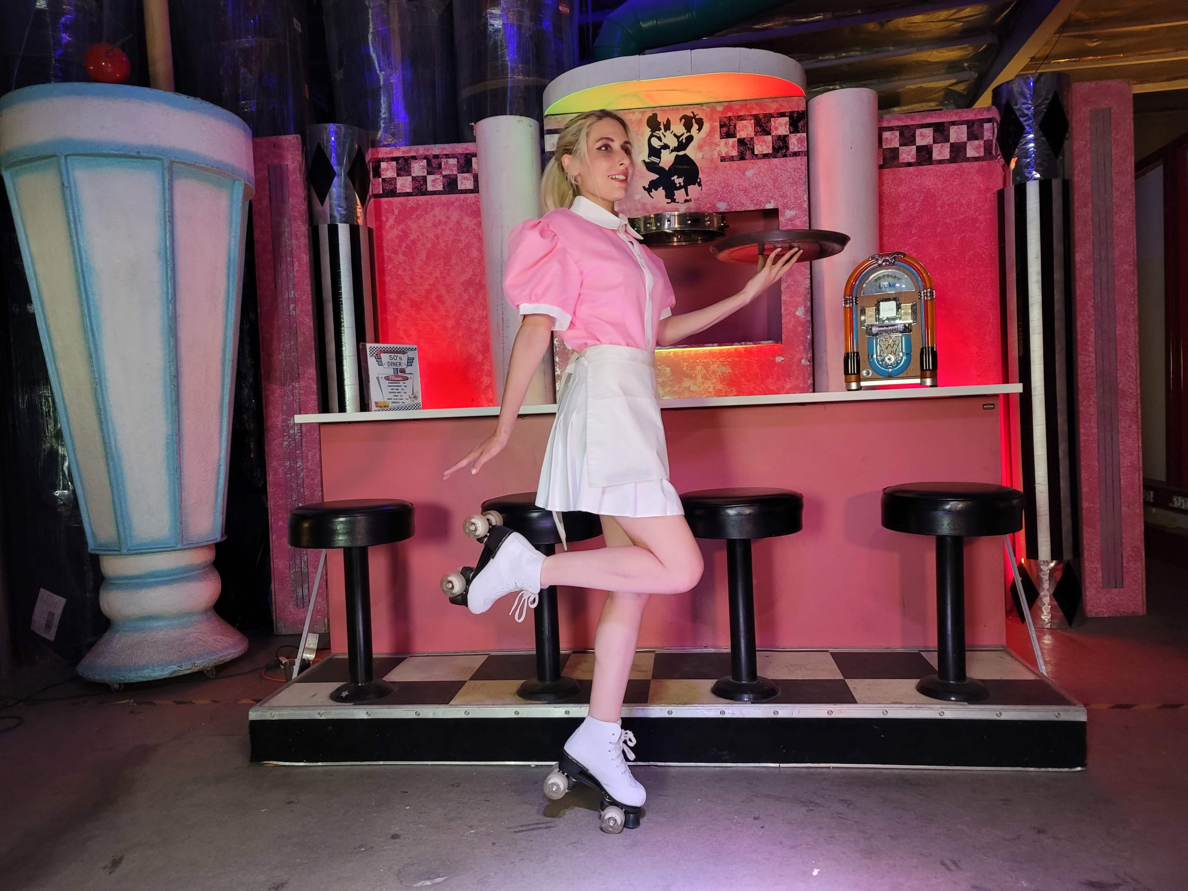 A woman on roller skates poses with a tray in front of a vintage diner-style bar with pink decor and black stools.