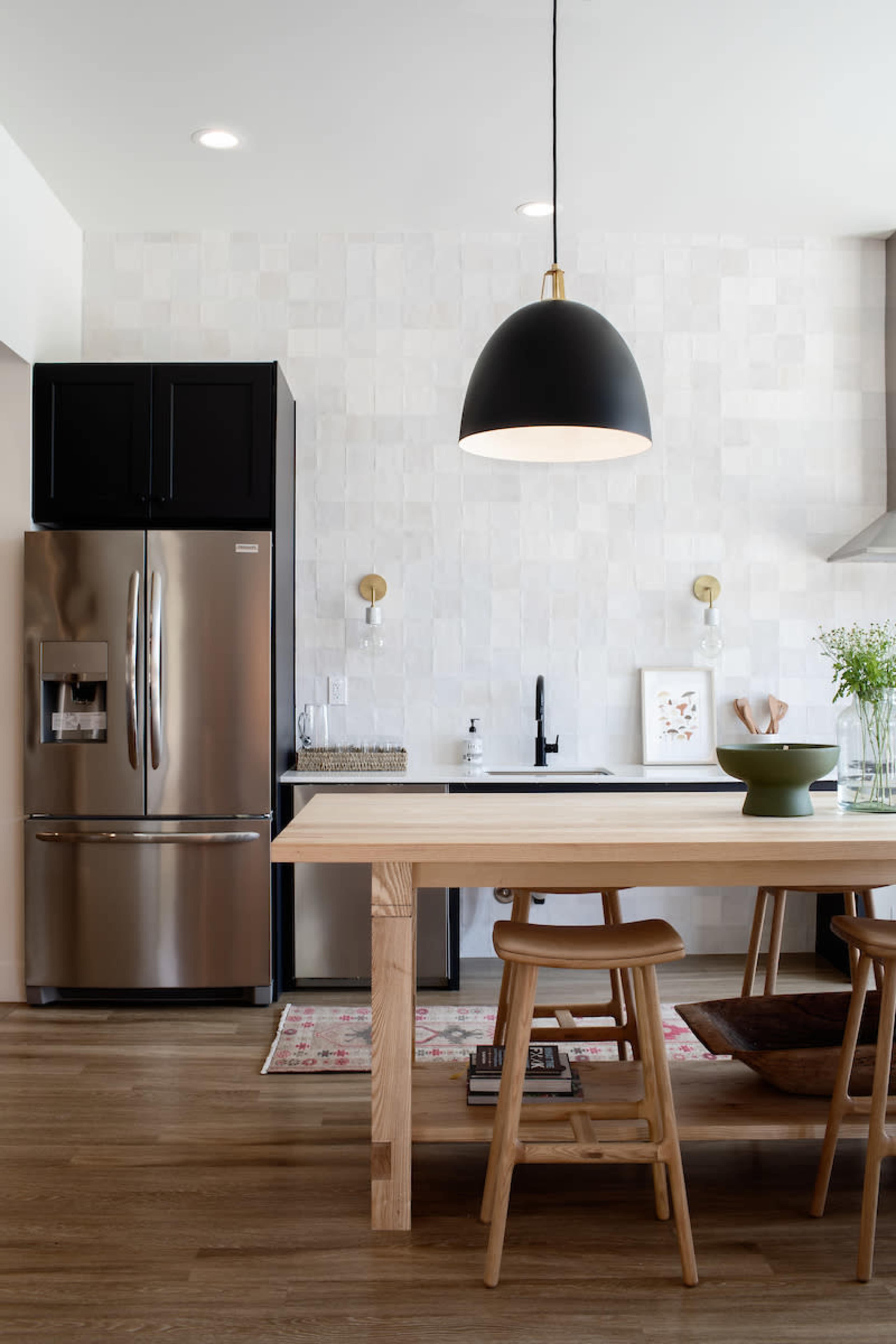 The image shows a modern kitchen with a wooden table and chairs, a stainless steel refrigerator, and a light fixture hanging above.
