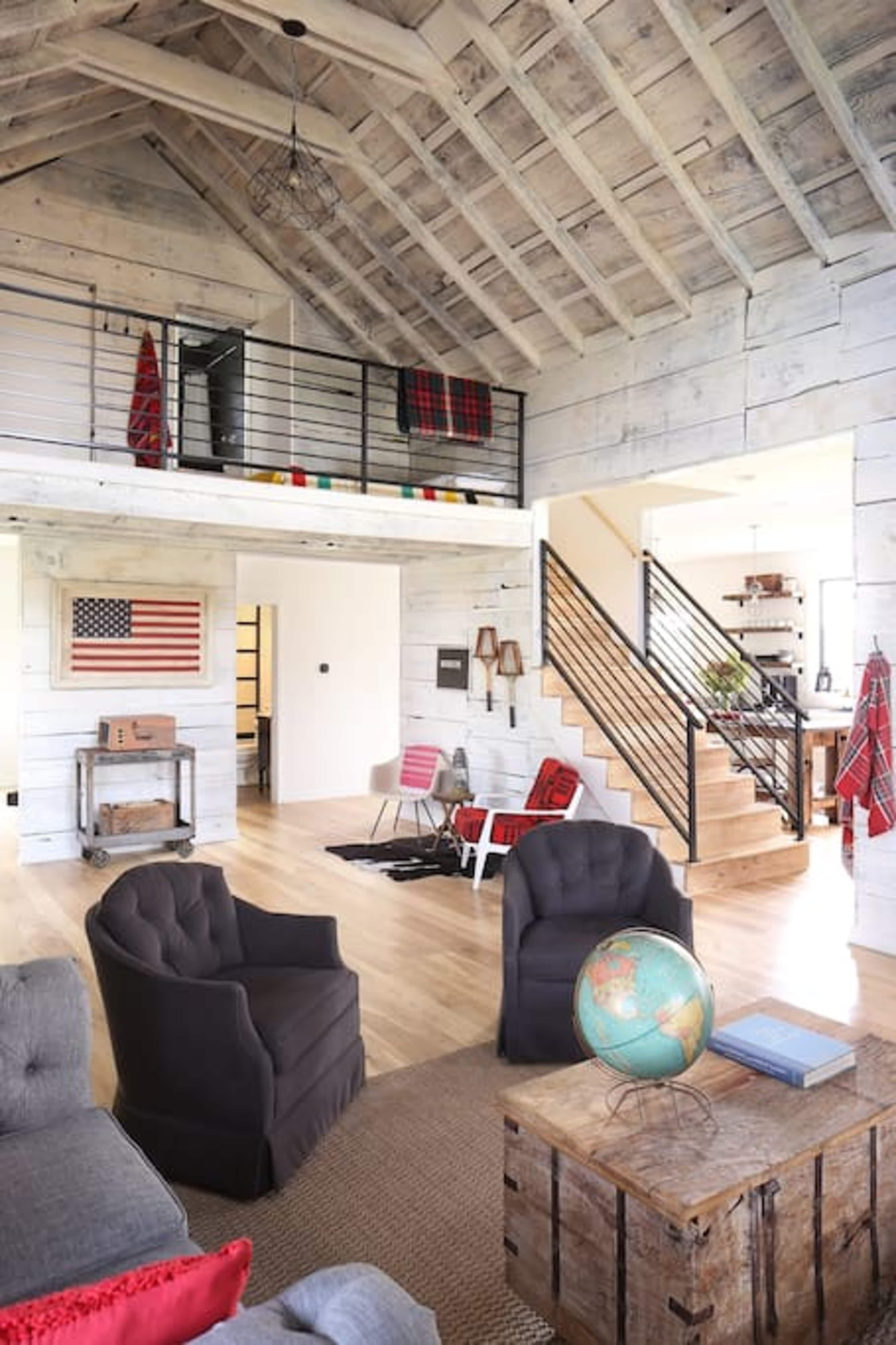 A spacious, modern living room with a wooden ceiling, a staircase, and comfortable seating arrangements, featuring an American flag on the wall and a globe on a coffee table.