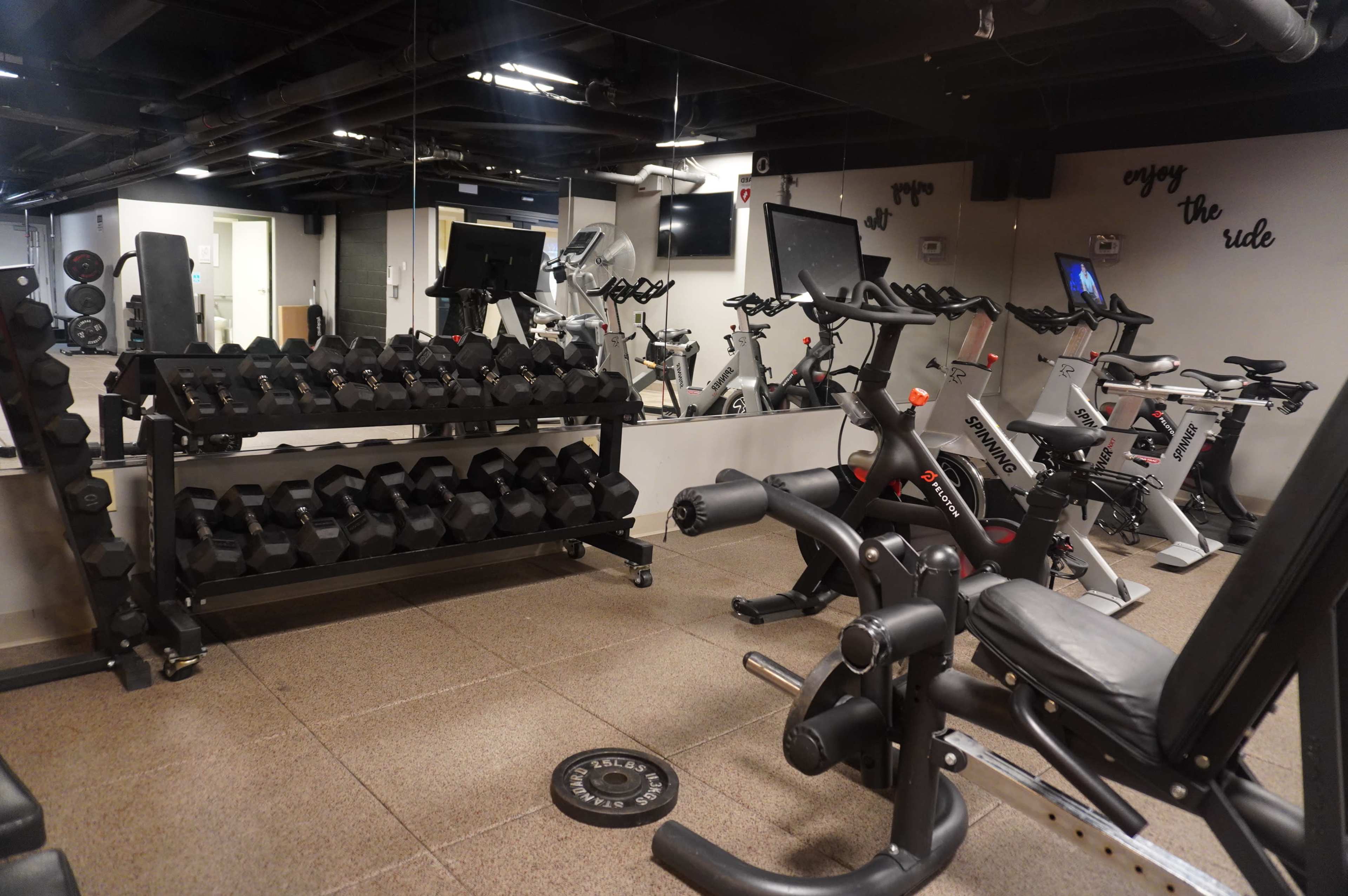 The image shows a gym with a rack of dumbbells, exercise bikes, and weight training equipment arranged in a well-lit space.