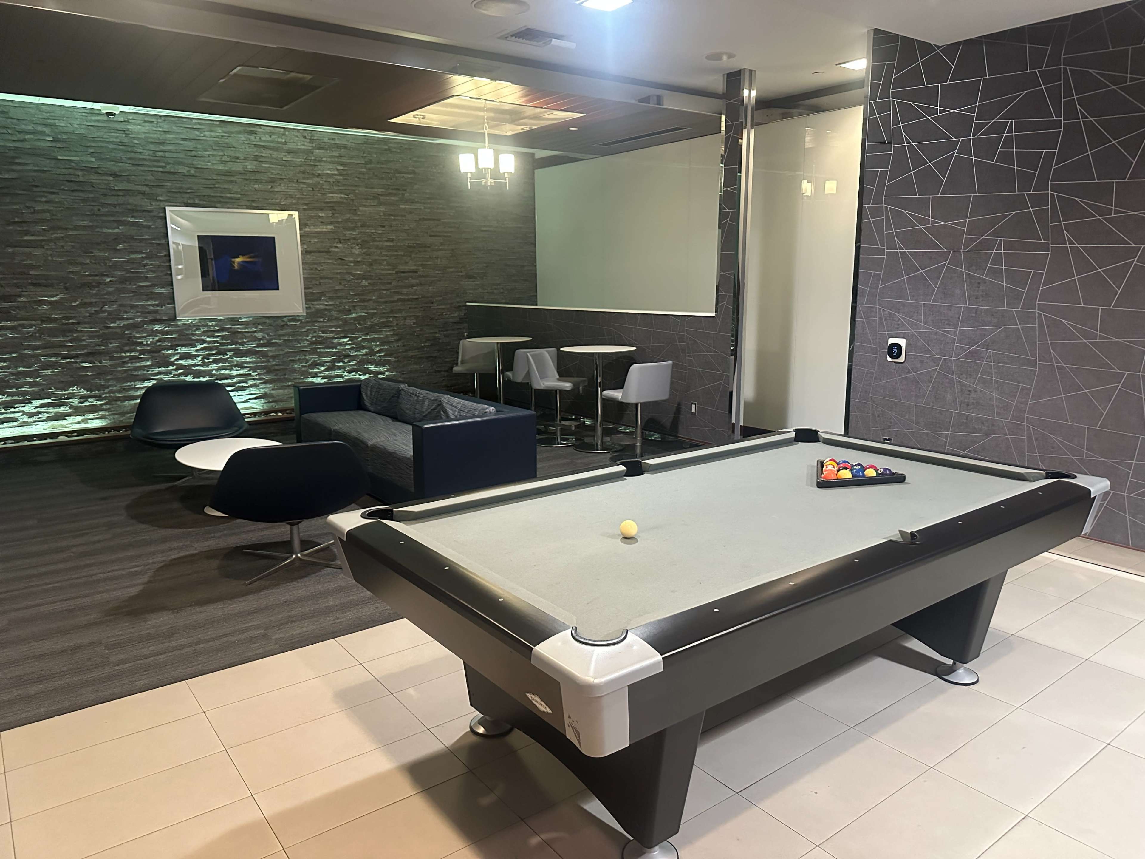 The image shows a modern lounge area featuring a pool table, a seating arrangement with a couch and chairs, and soft lighting against a textured wall.