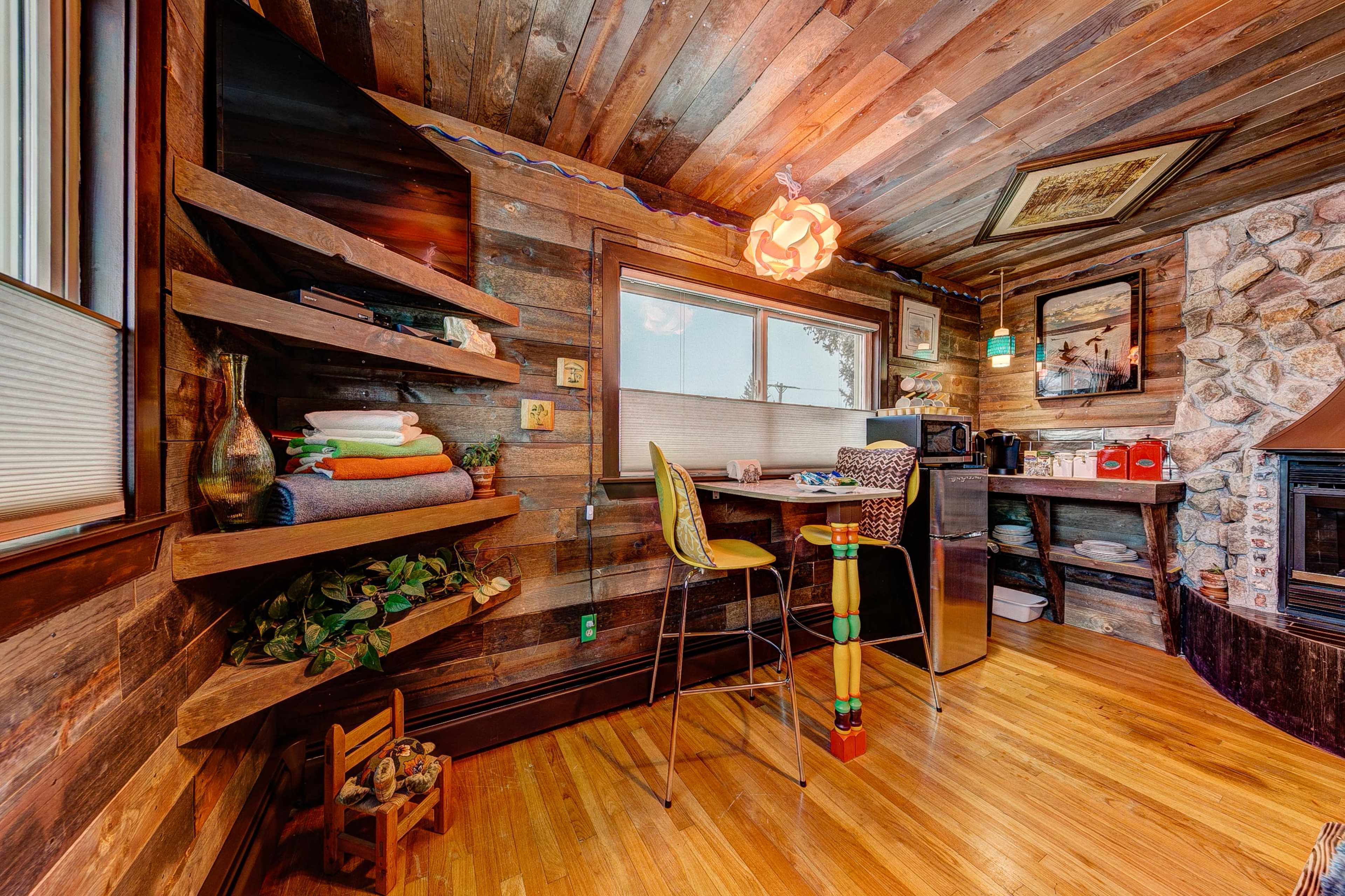 A cozy wooden room featuring a small kitchenette with shelves, a microwave, and a bar stool next to a stone fireplace.