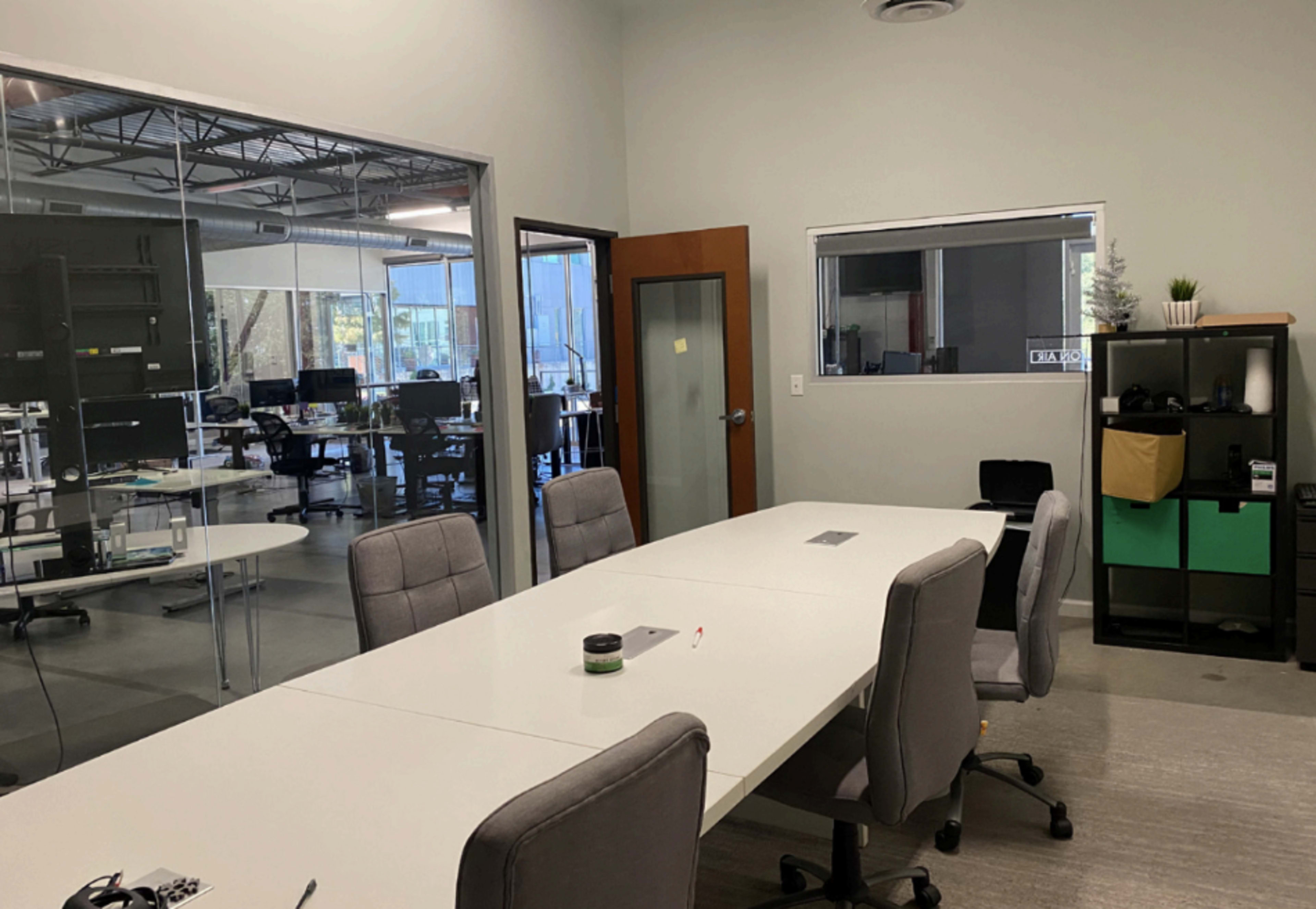 A modern office space features a large conference table surrounded by gray chairs, with a glass wall revealing additional workstations in the background.