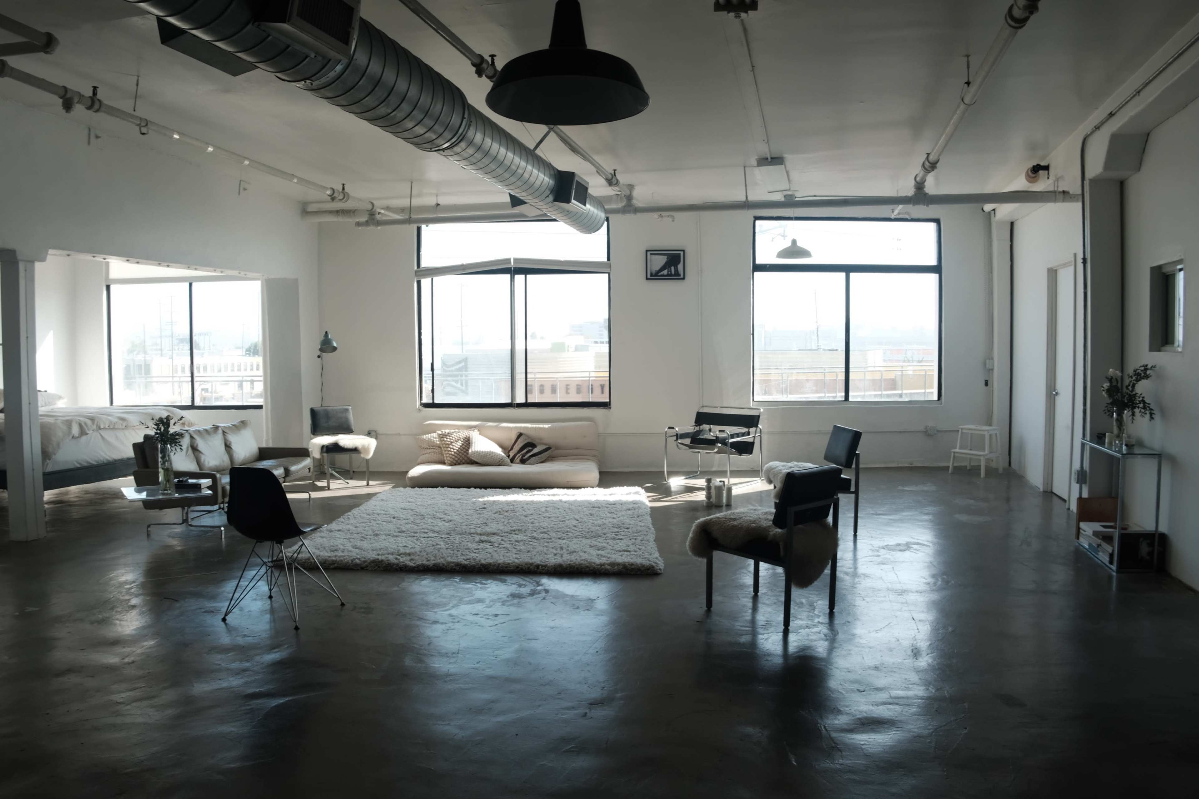 The room features a spacious, modern loft with large windows, minimalist furniture, and a polished concrete floor.