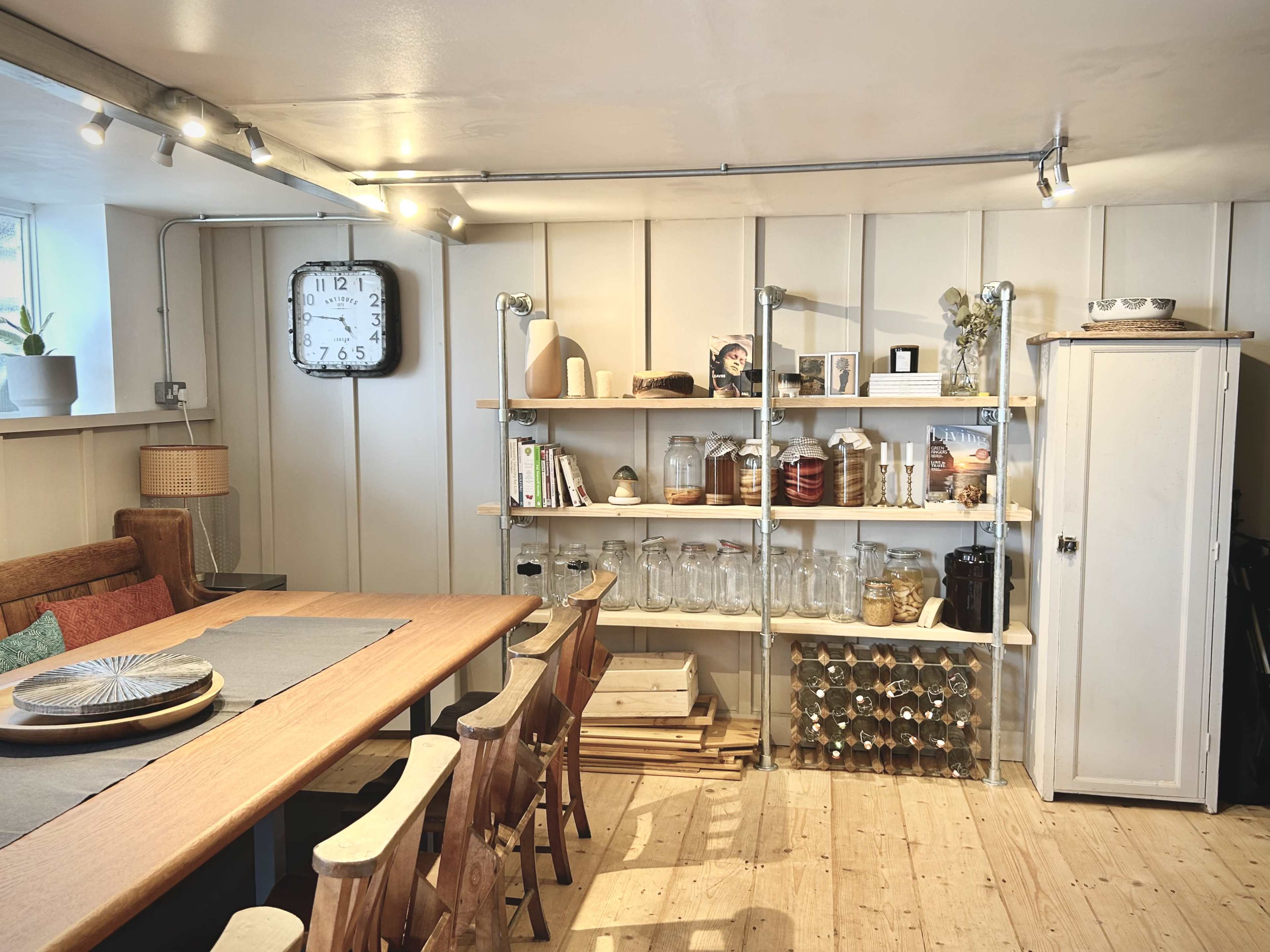 A dining area features a large table surrounded by wooden chairs, with shelves displaying various jars and objects along one wall.