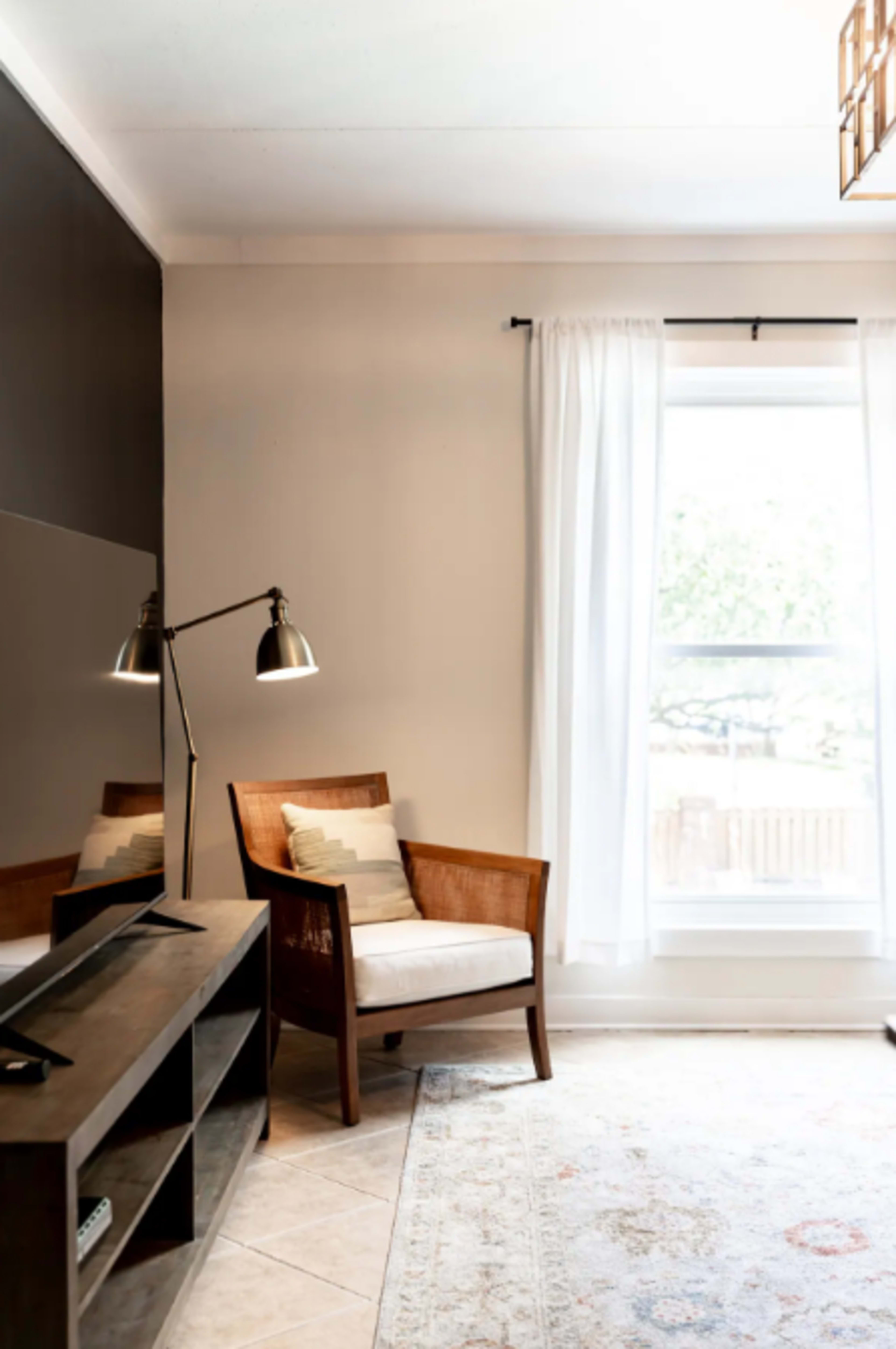 A simple living room featuring a wooden chair with a cushion, a floor lamp, a TV stand, and a large window with sheer curtains.