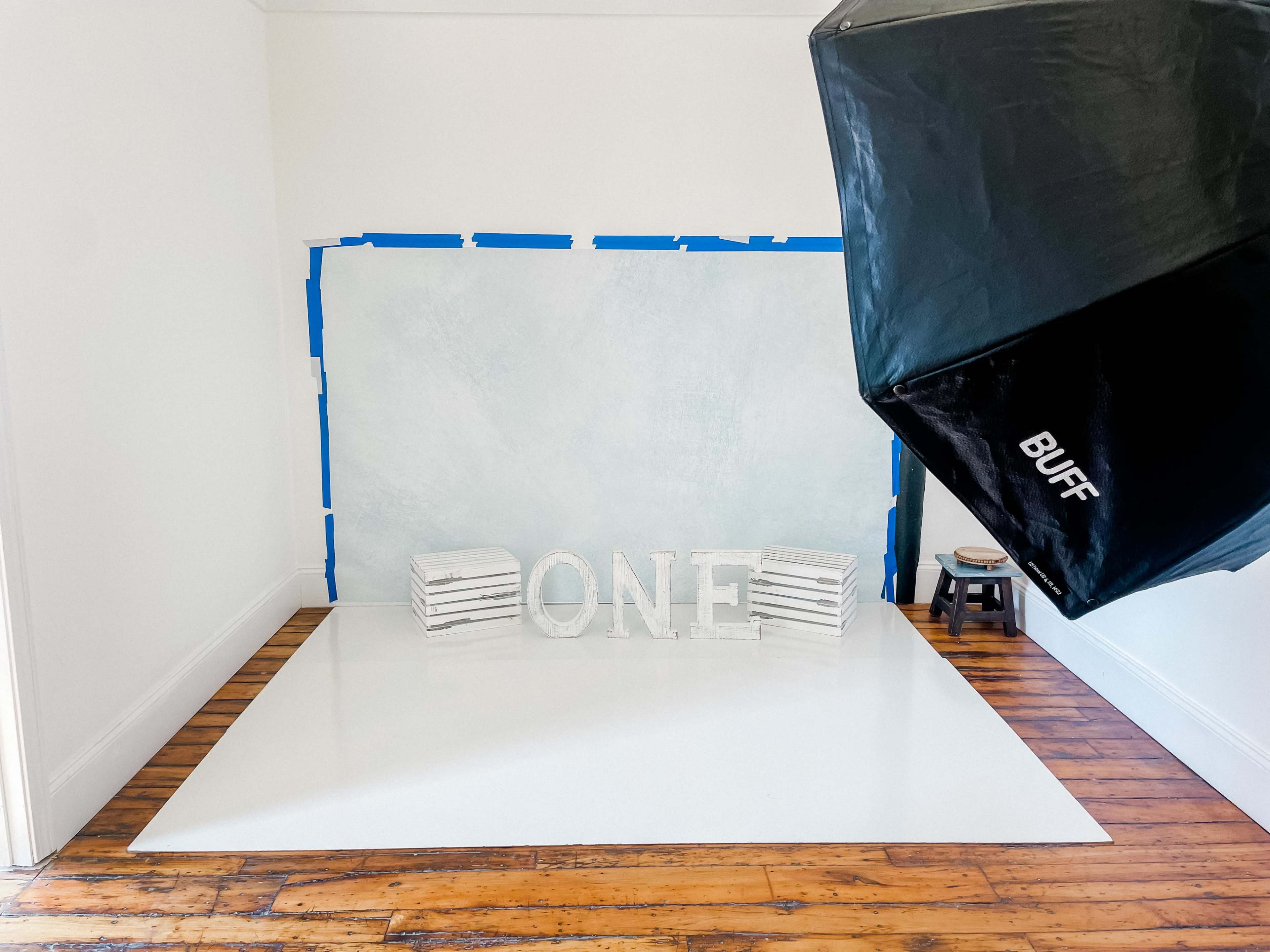A spacious, well-lit photography studio features a blue-taped backdrop, white floor, and a large, decorative "ONE" displayed in the center.