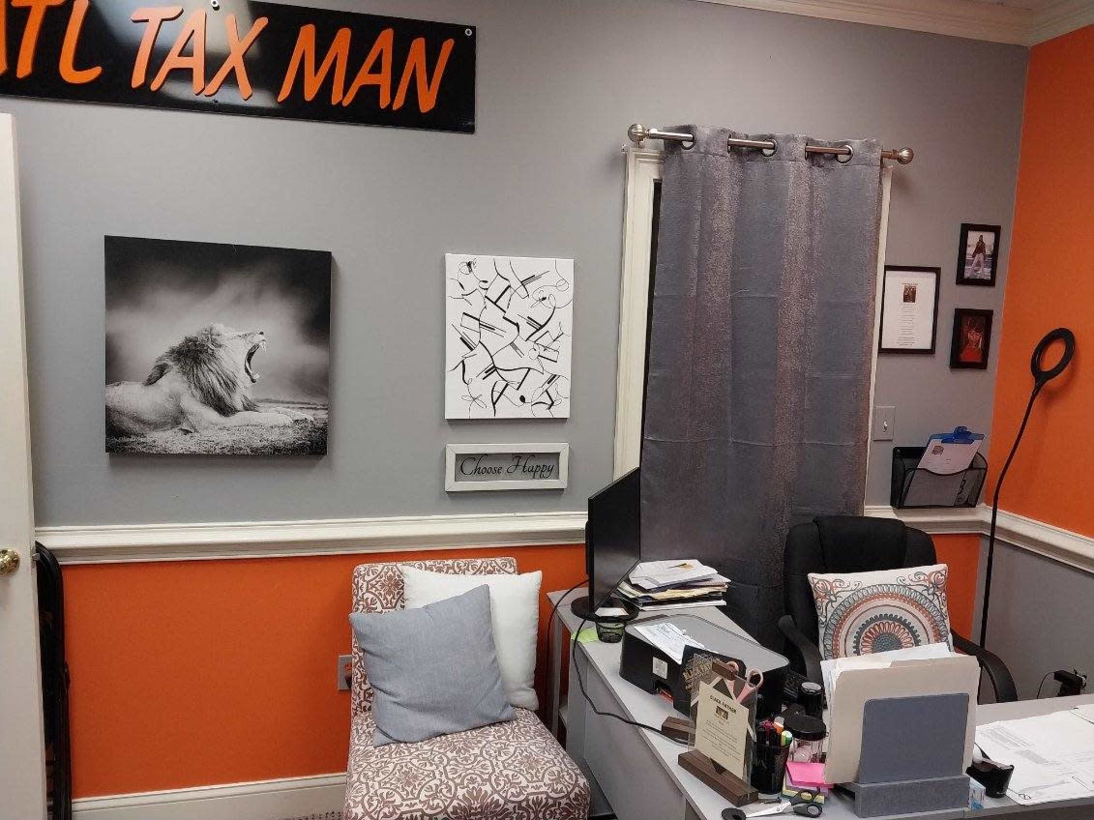 An office space with orange and gray walls featuring a lion photograph and abstract art, a cluttered desk with an open laptop, and a chair with decorative cushions.