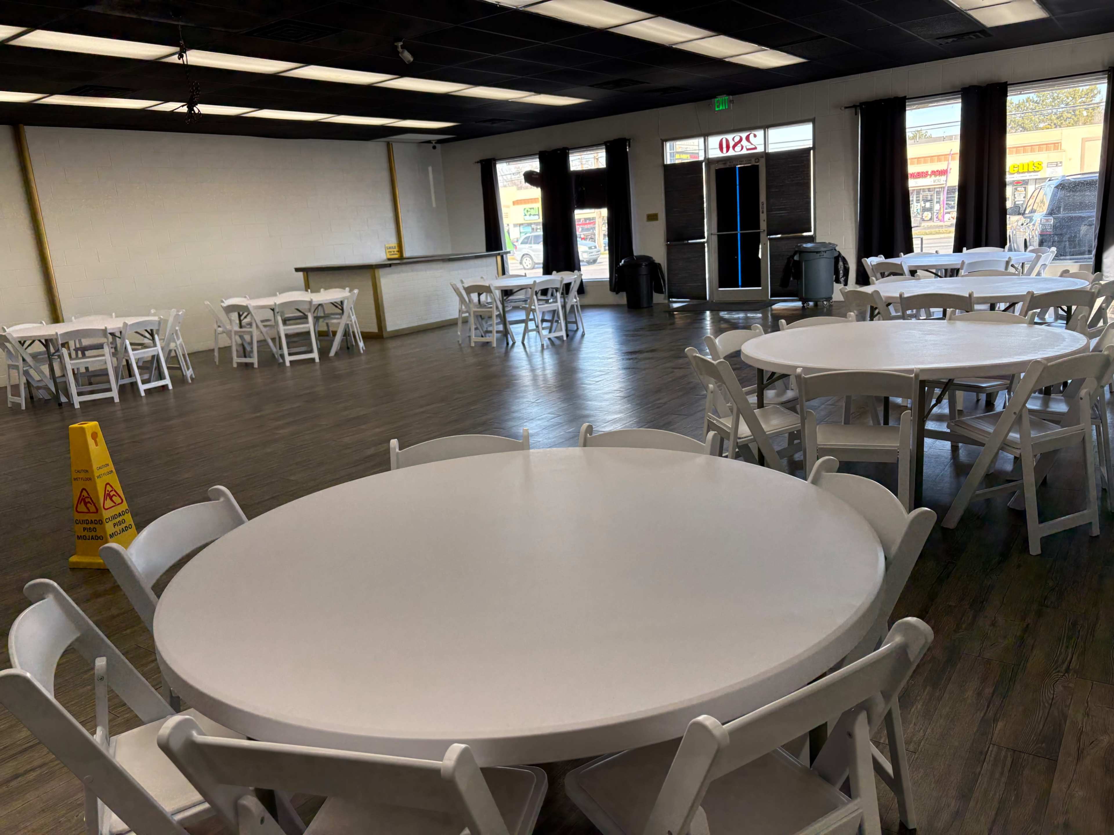 The image shows an empty event space with white round tables and chairs arranged around the room, featuring a large entrance door and a caution sign on the floor.