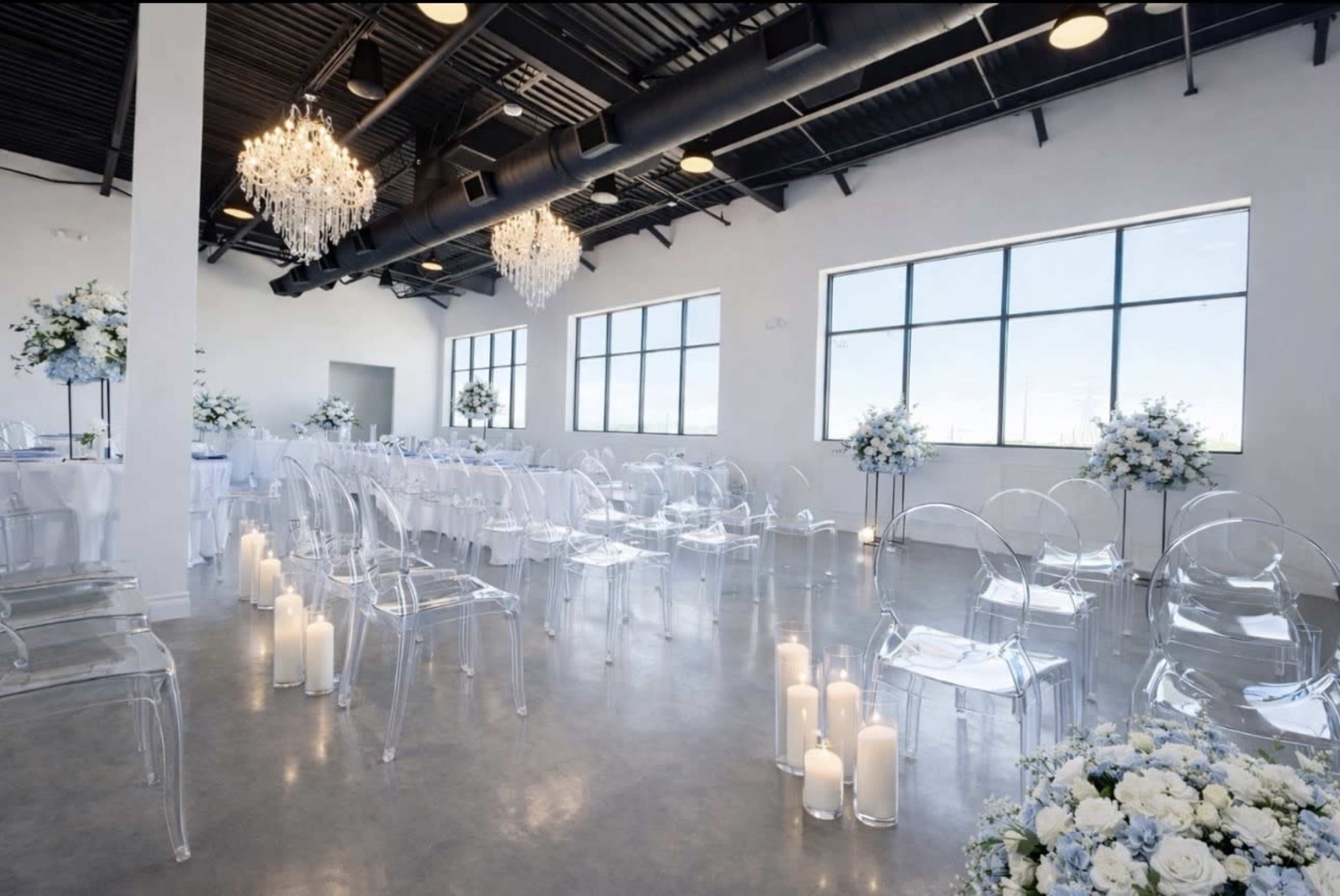 A spacious event venue features rows of clear chairs, elegant floral arrangements, and candles lining the floor beneath large windows.