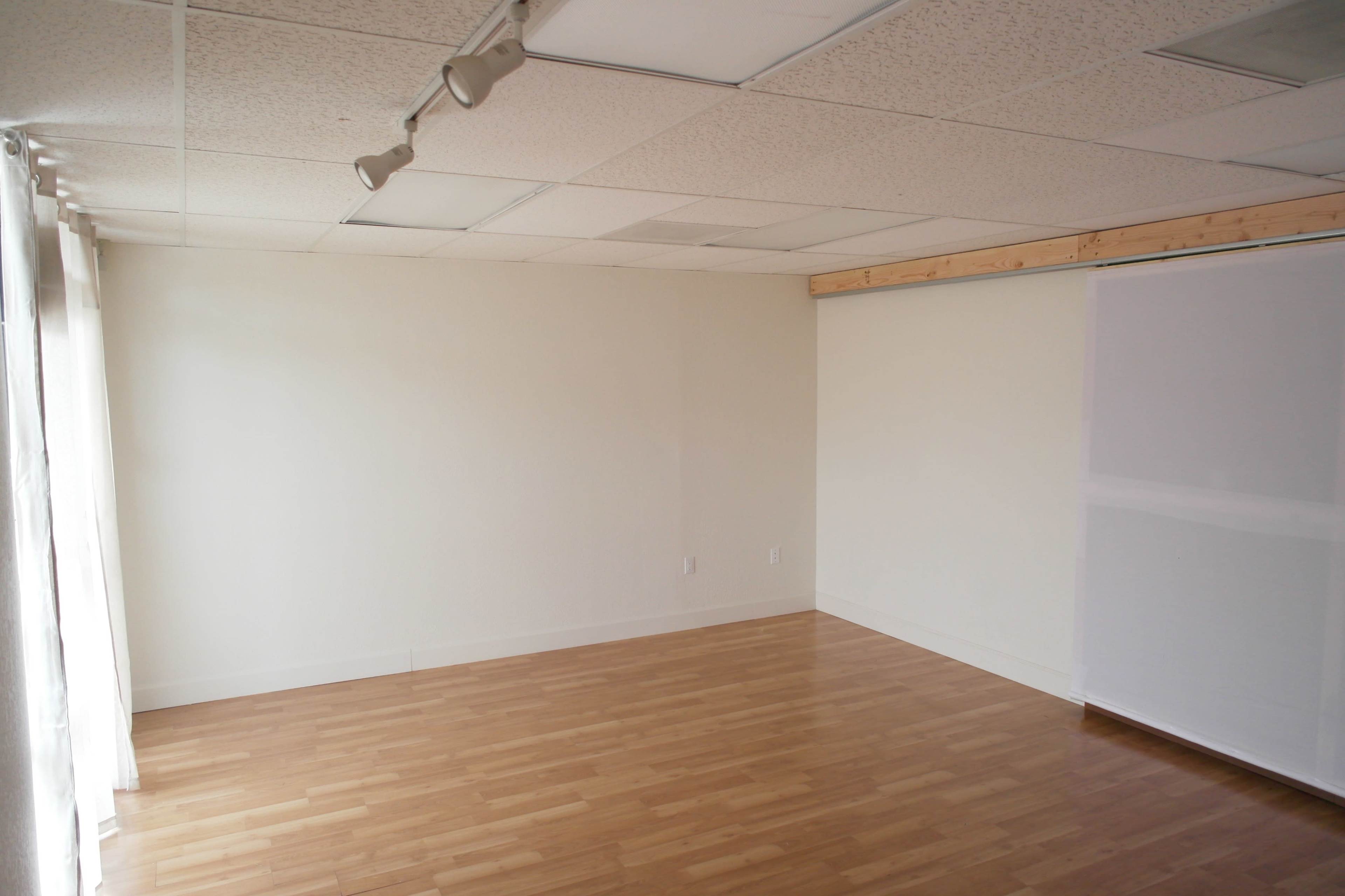 An empty room with light wood flooring, white walls, and a window covered by a sheer curtain.