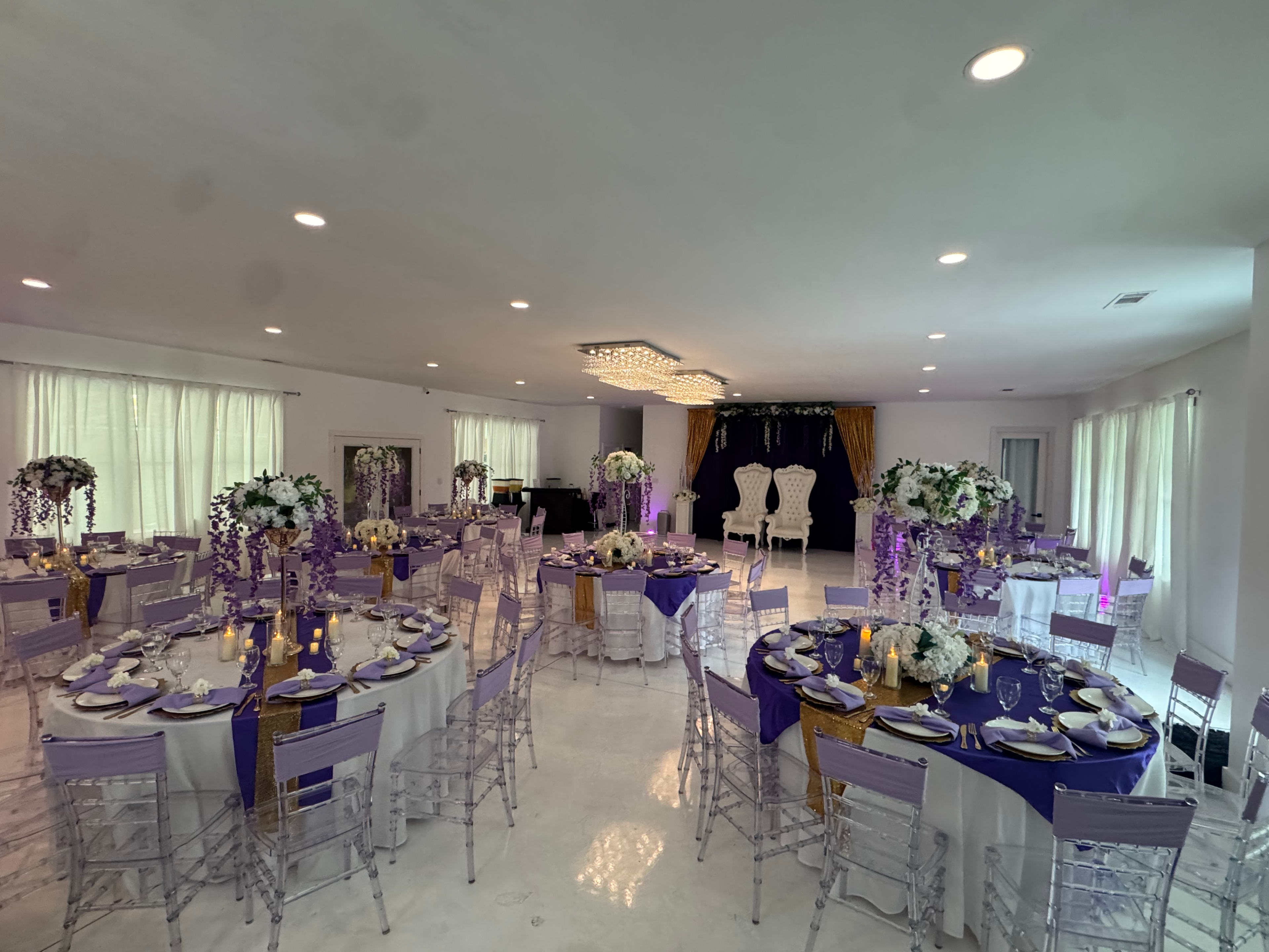 The image shows an elegantly set banquet hall with round tables dressed in white and purple, adorned with floral centerpieces and candles, and a decorative stage in the background.