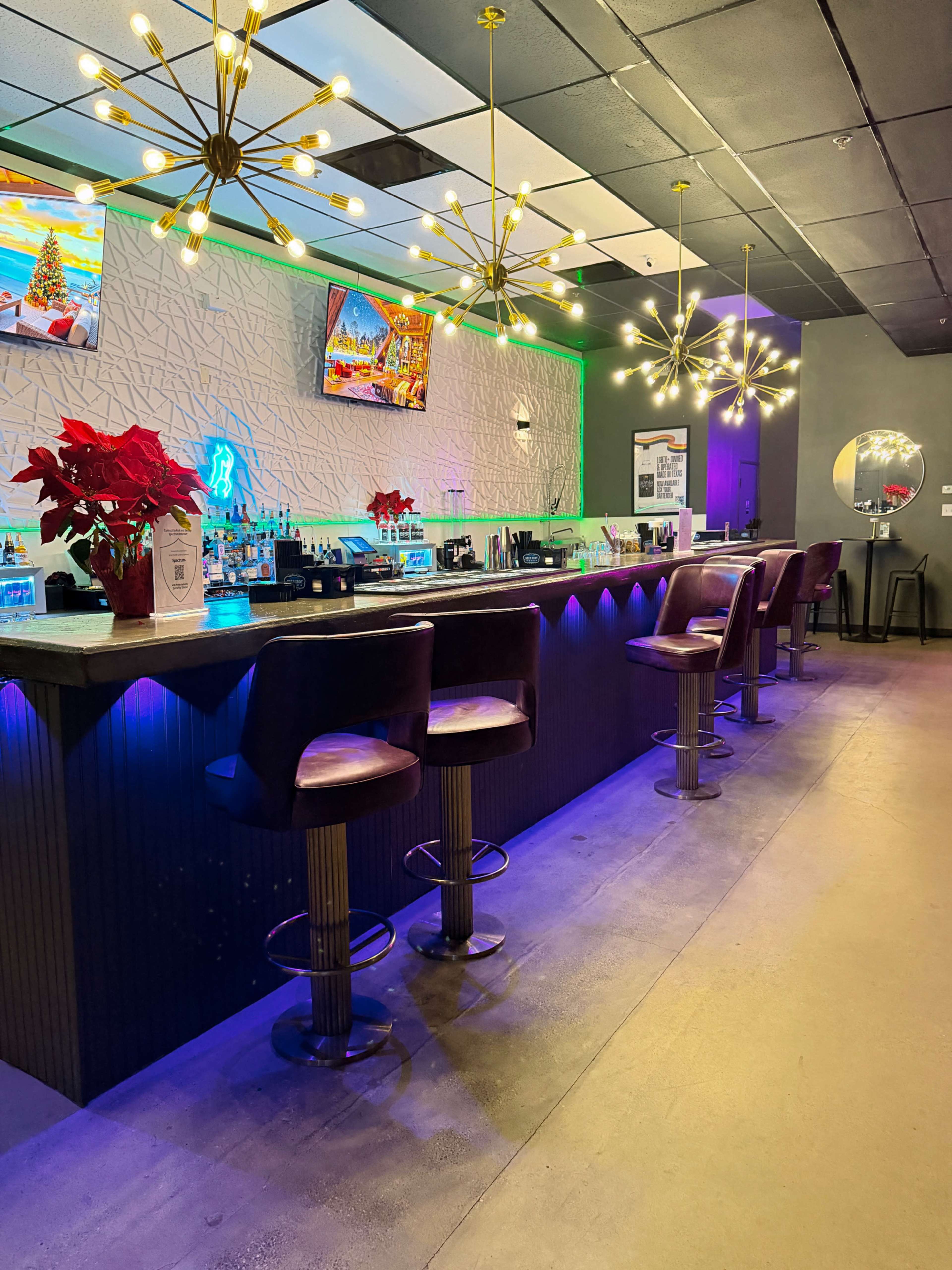 The image shows a modern bar area with illuminated shelves, stylish bar stools, and decorative lighting fixtures, featuring a festive touch with poinsettias on the counter.