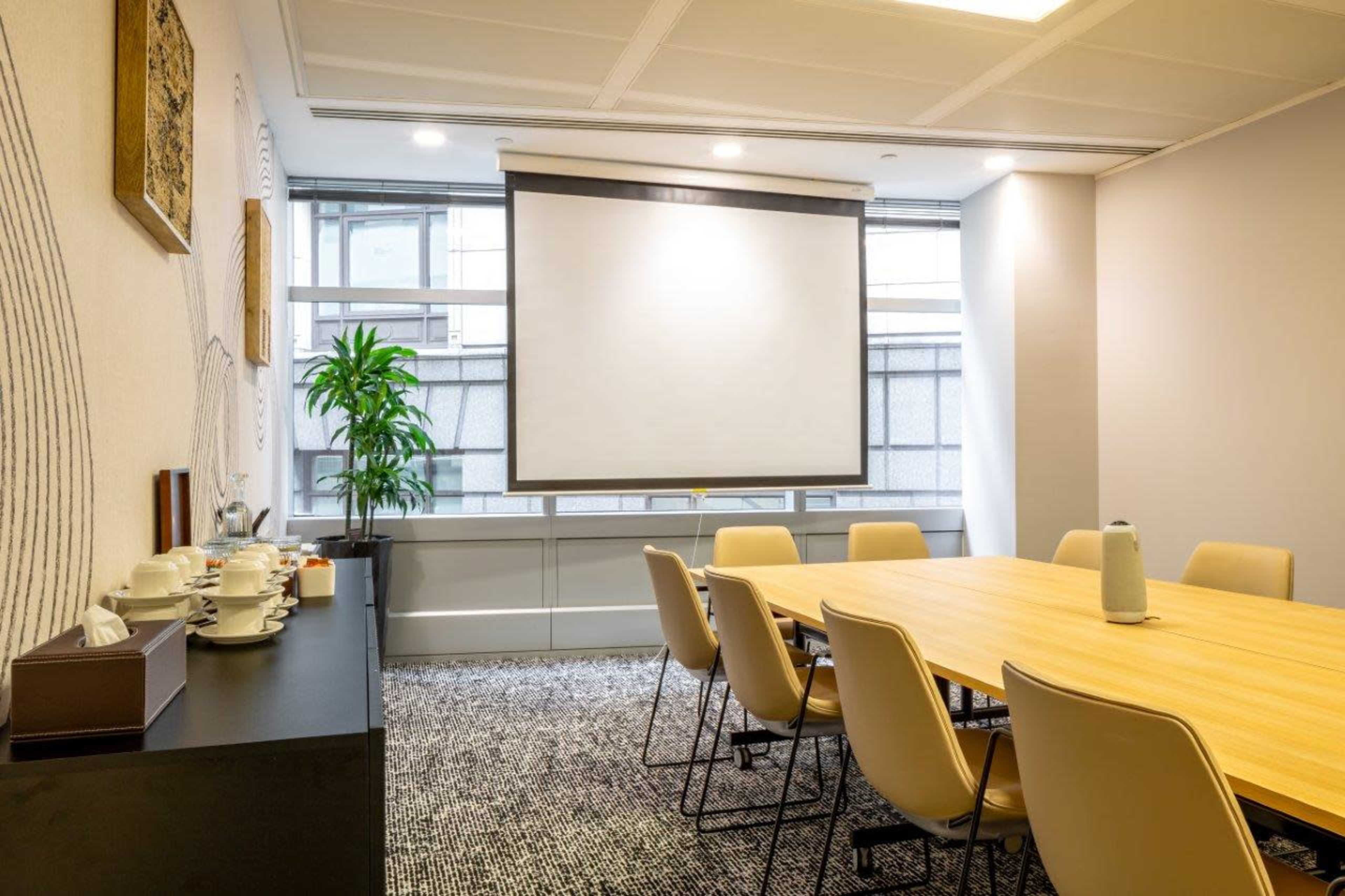 A conference room features a large wooden table surrounded by beige chairs, with a projector screen on one wall and a plant in the corner.