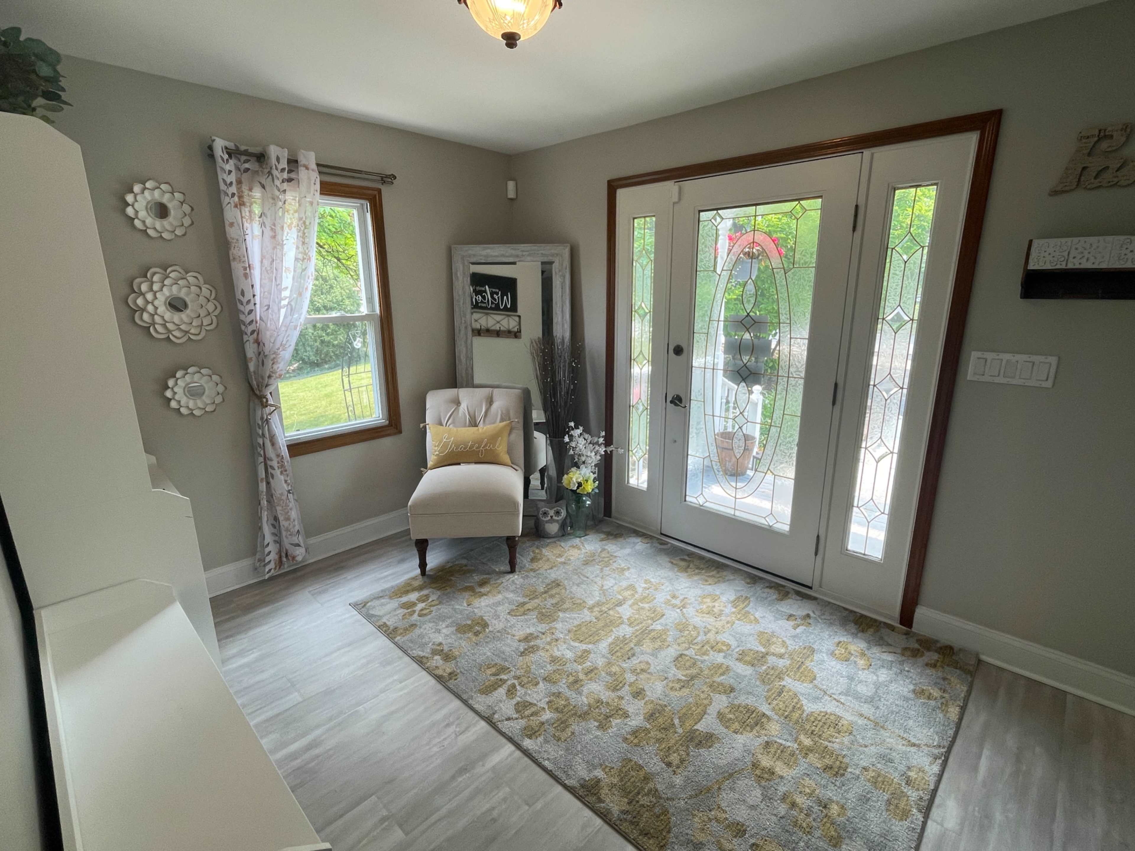 The image shows a well-lit entryway featuring a chair, a decorative rug, a mirror, and a door with a glass panel.