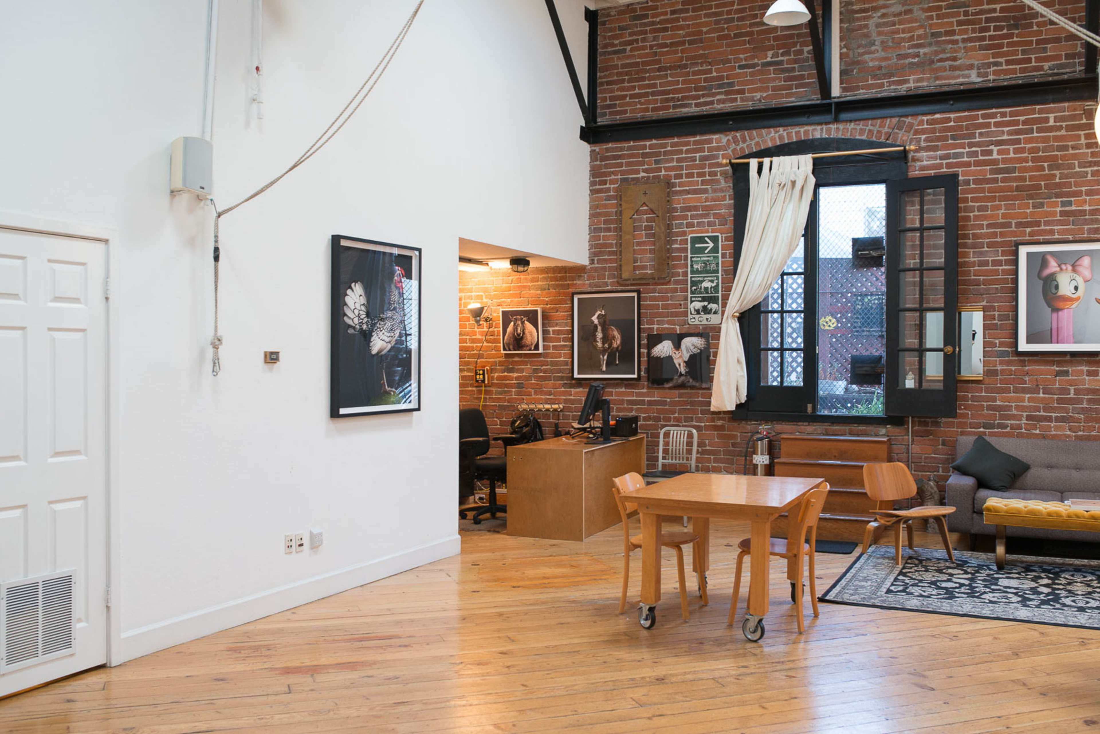 The image shows an open loft space with exposed brick walls, a wooden floor, a table and chairs, and a workspace featuring artwork on the walls.