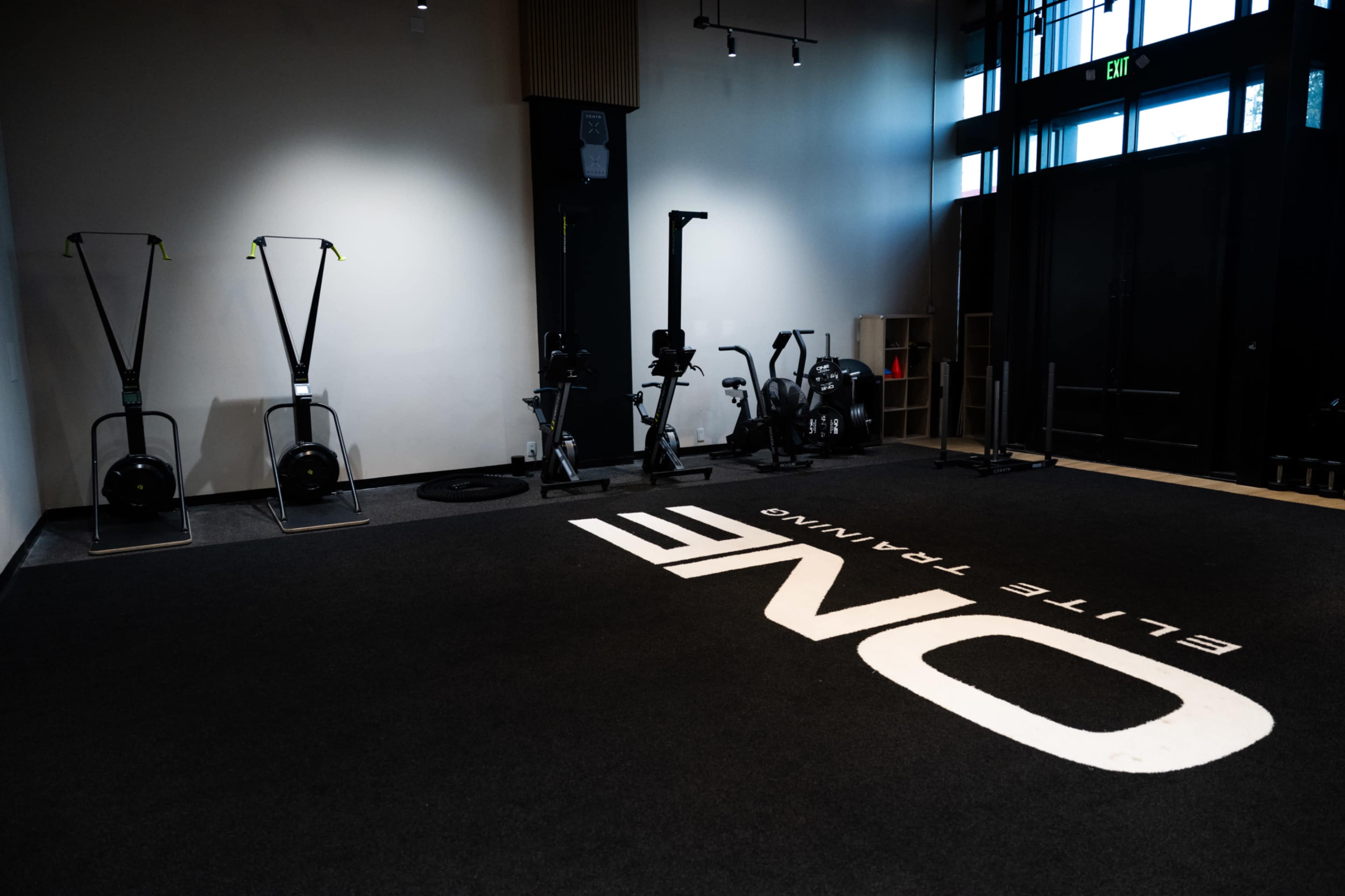 The image shows a fitness training space featuring various equipment, including rowers and stationary bikes, with "ONE ELITE TRAINING" prominently displayed on the floor.