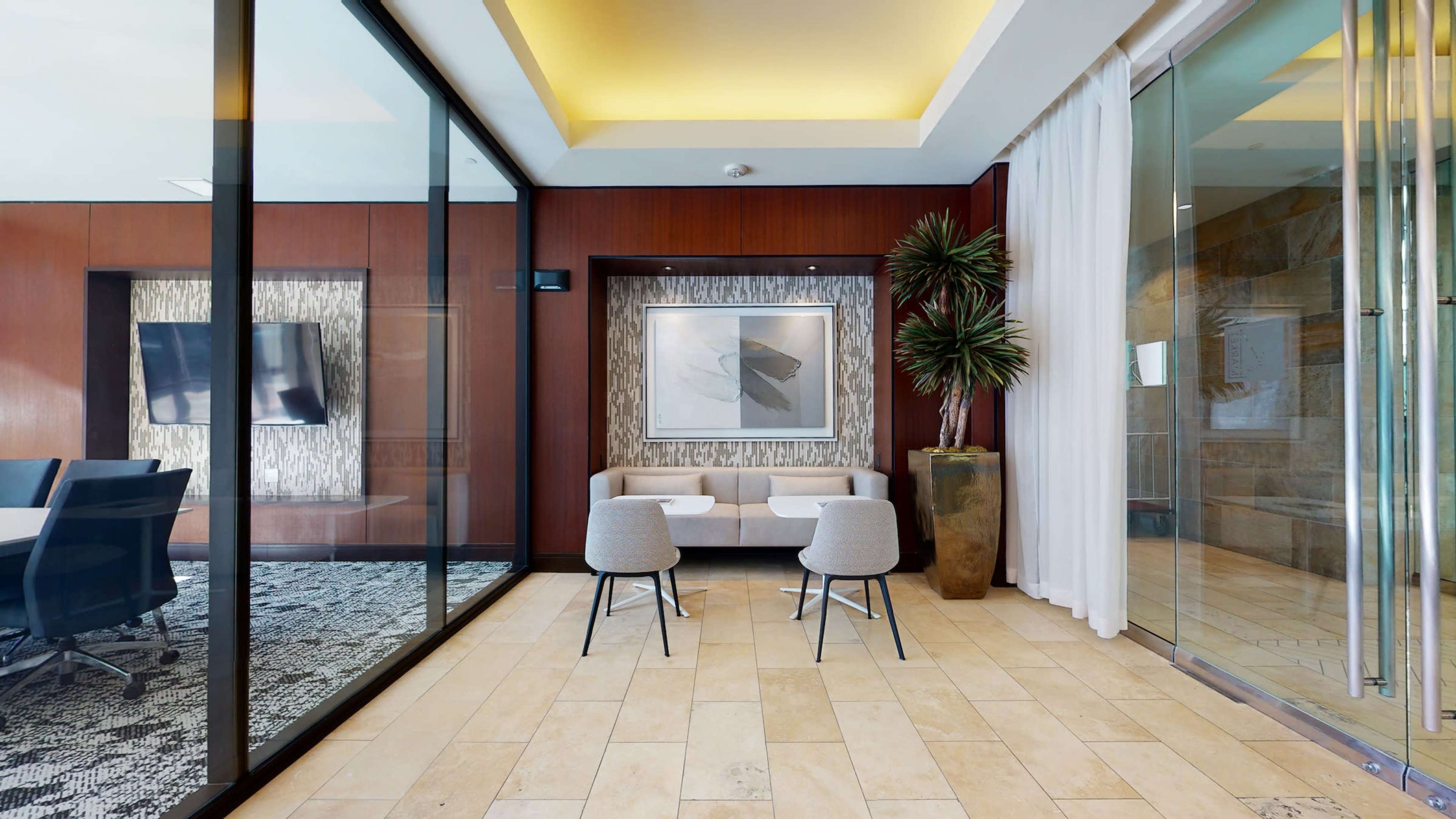 A modern meeting space features a glass wall, two chairs facing a small table, and decorative elements including a potted plant and wall art.