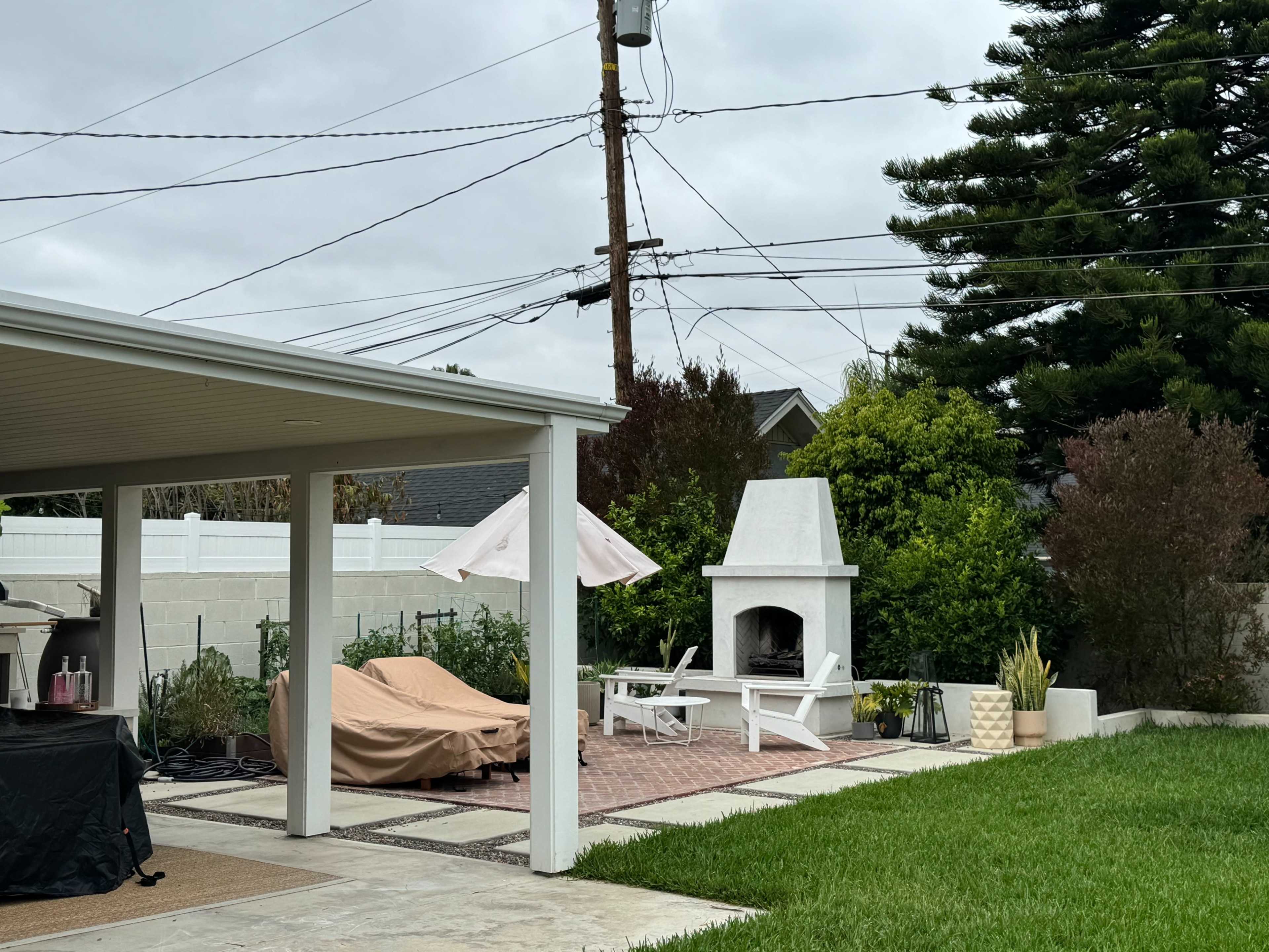 The image shows a backyard with a covered patio, a brick fireplace, outdoor seating, and power lines overhead.