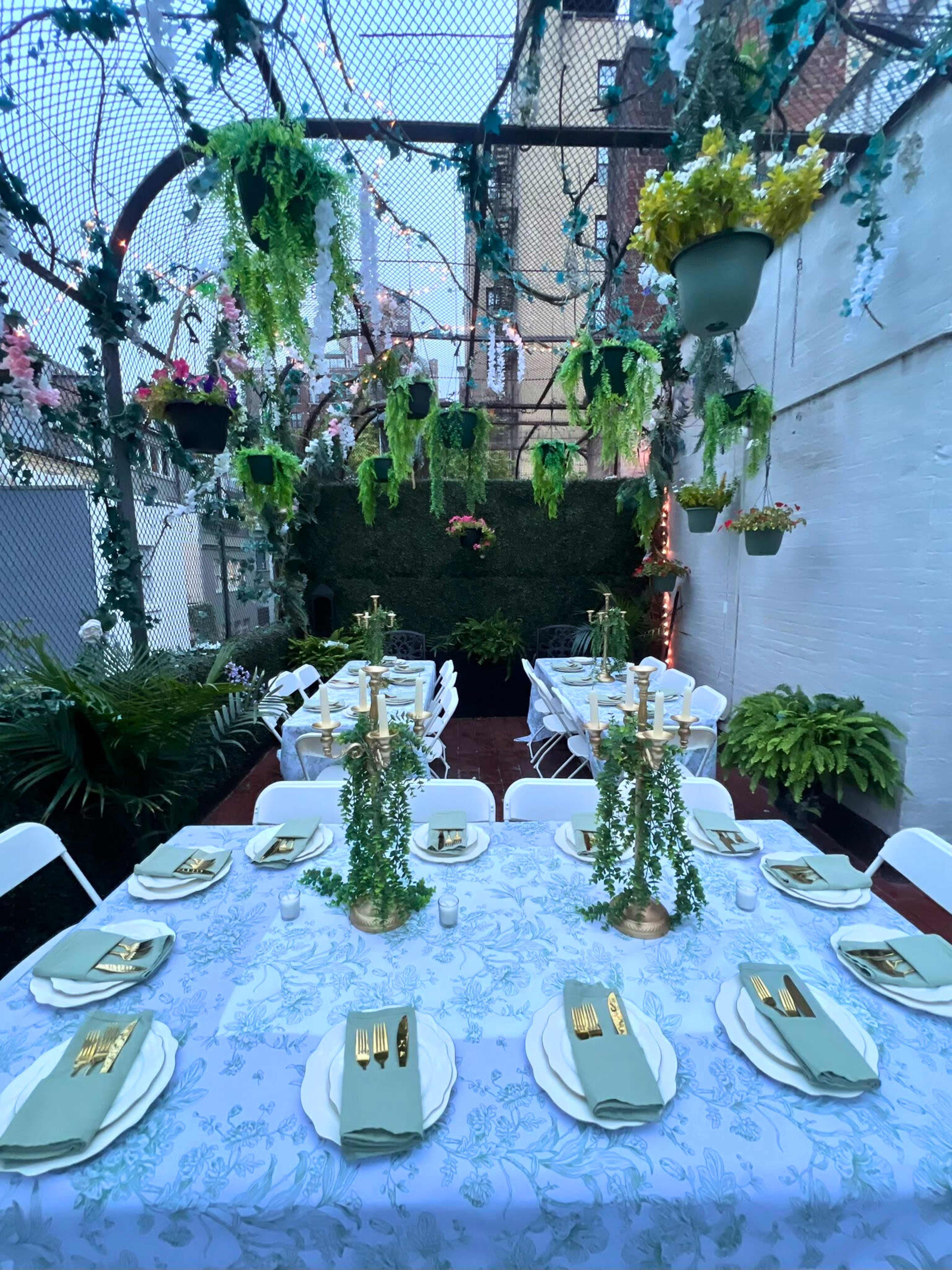 A decorated outdoor dining area features a long table set with gold utensils and green napkins, surrounded by hanging plants and fairy lights.