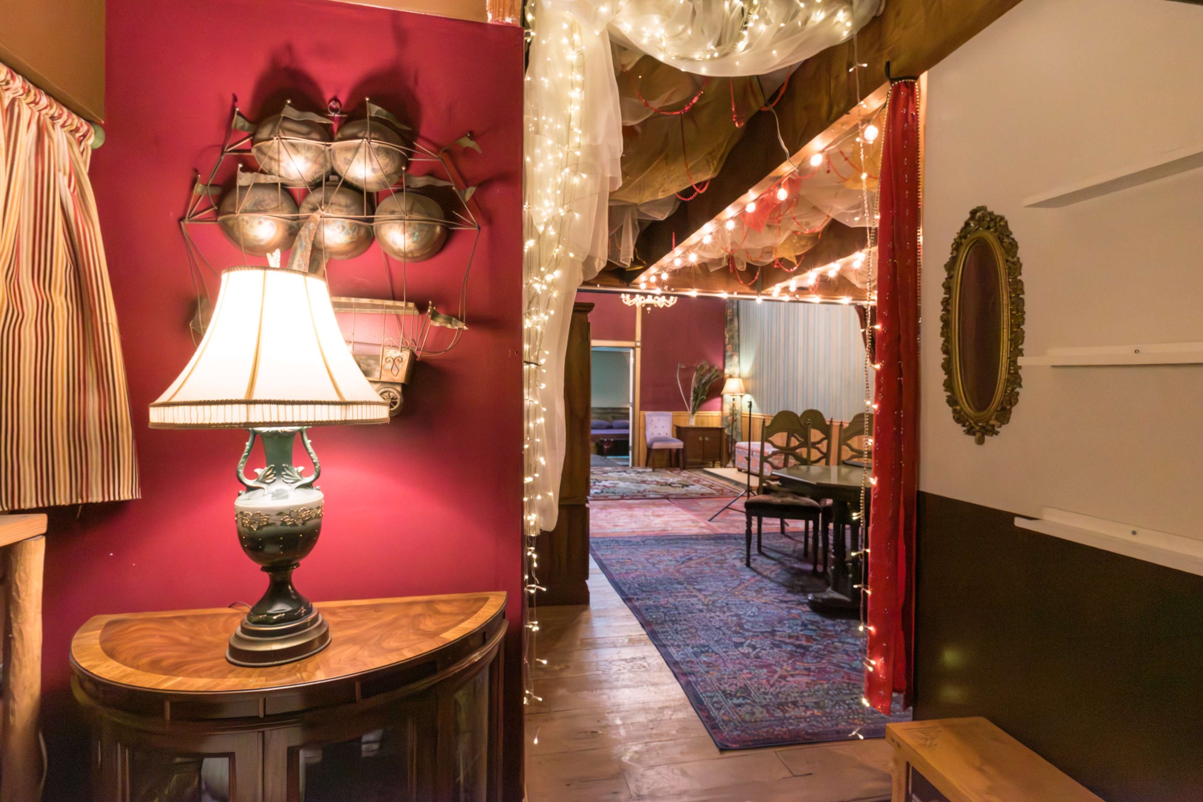 A warmly lit hallway adorned with string lights, leading to a room with a rug and furniture, framed by red walls and decorative elements.